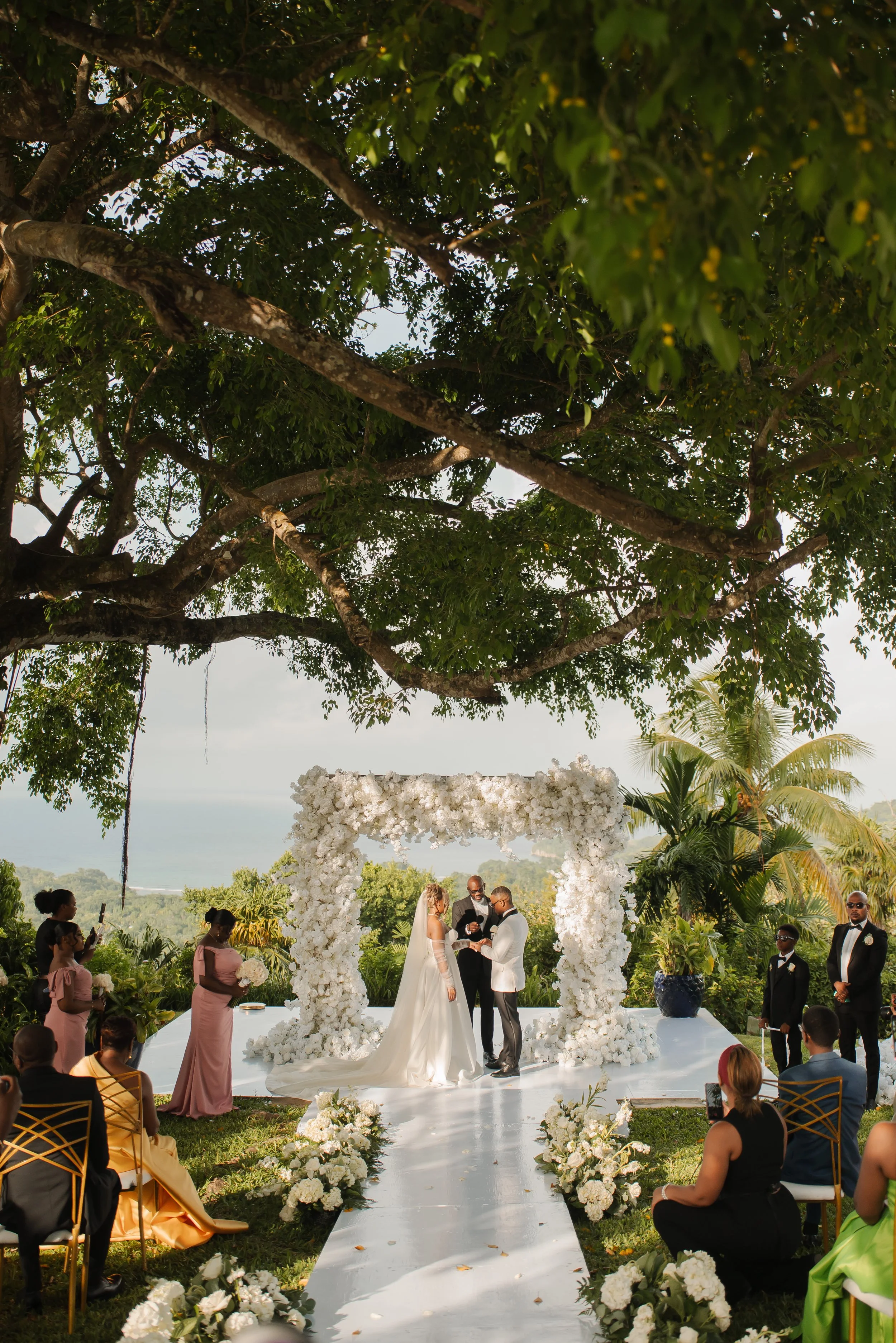 A wedding ceremony outdoors under a large tree with a floral arch, officiant, bride, and groom, surrounded by guests and lush greenery.