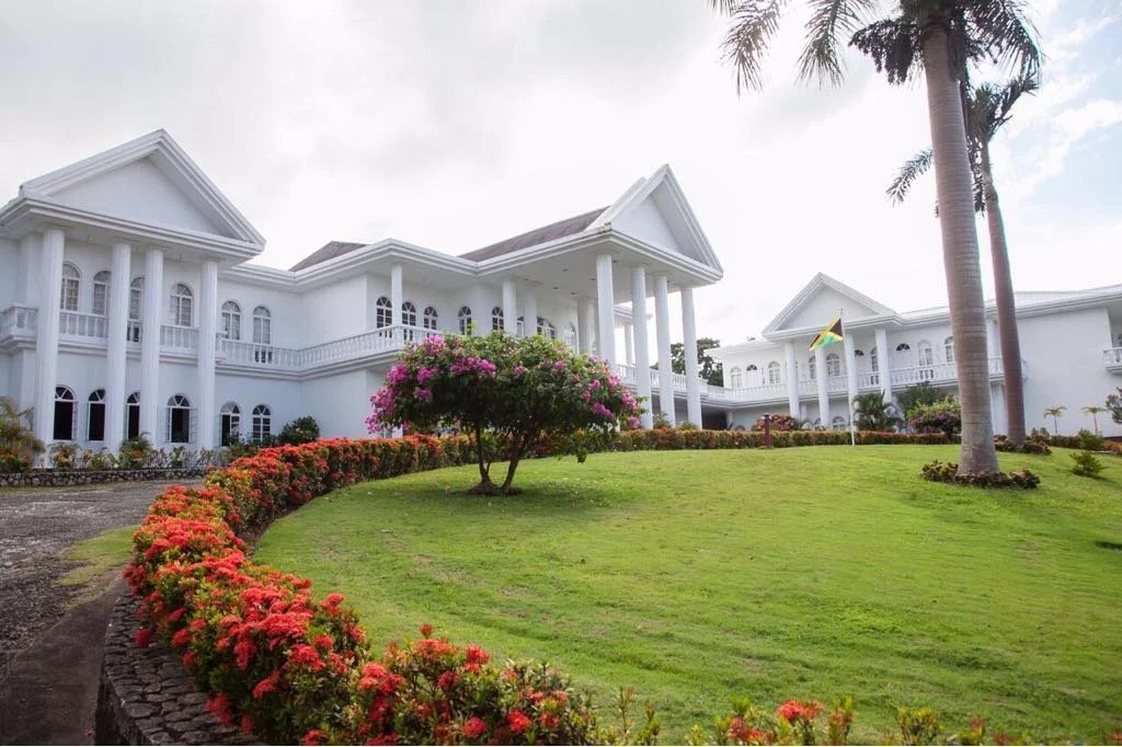 Large white mansion with columns, surrounded by well-maintained lawn, pink and red flowers, palm trees, and an island flag.