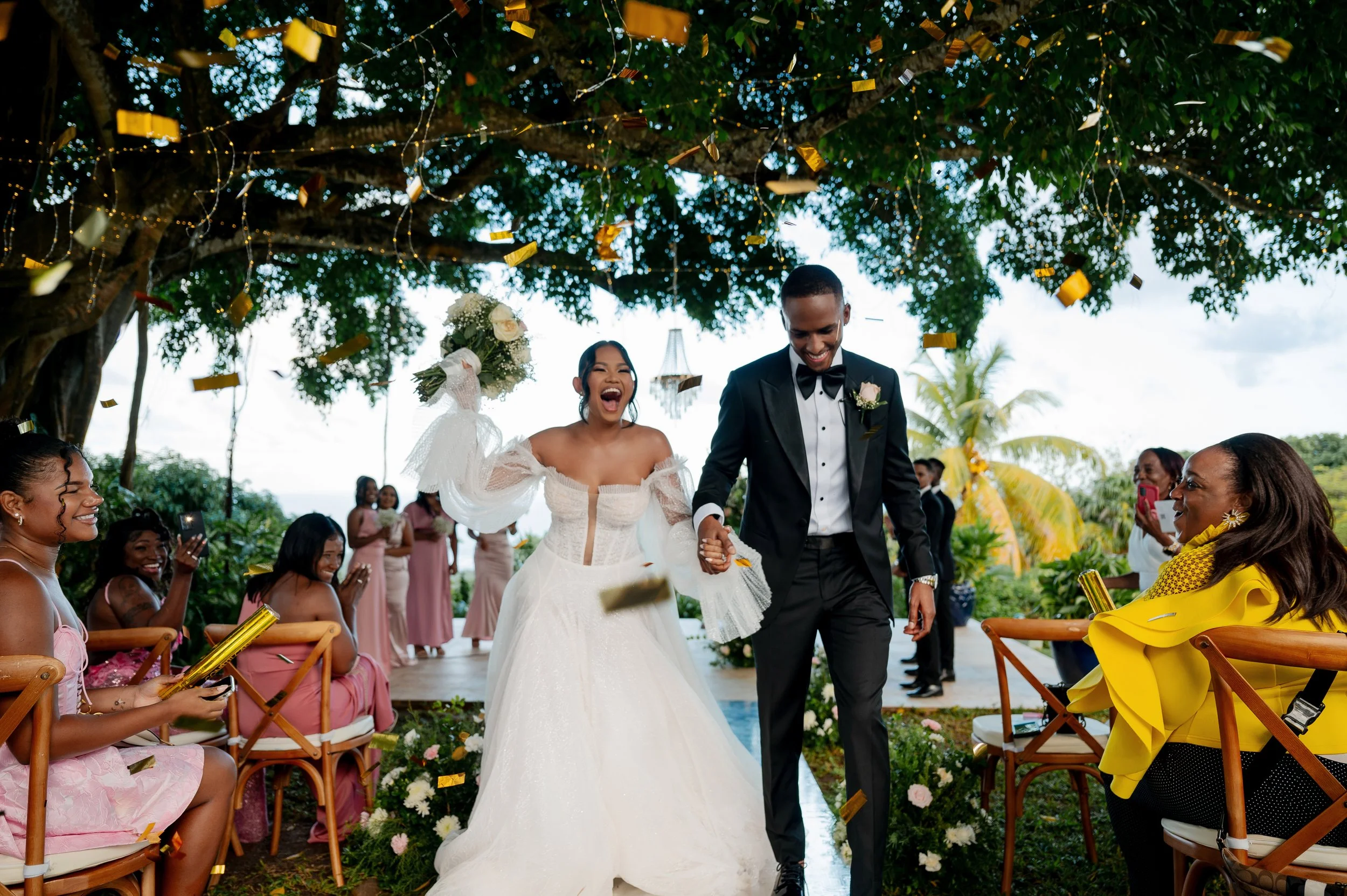 A wedding reception outdoors with a bride and groom walking down the aisle holding hands, surrounded by guests, greenery, and decorative lights, with confetti falling.