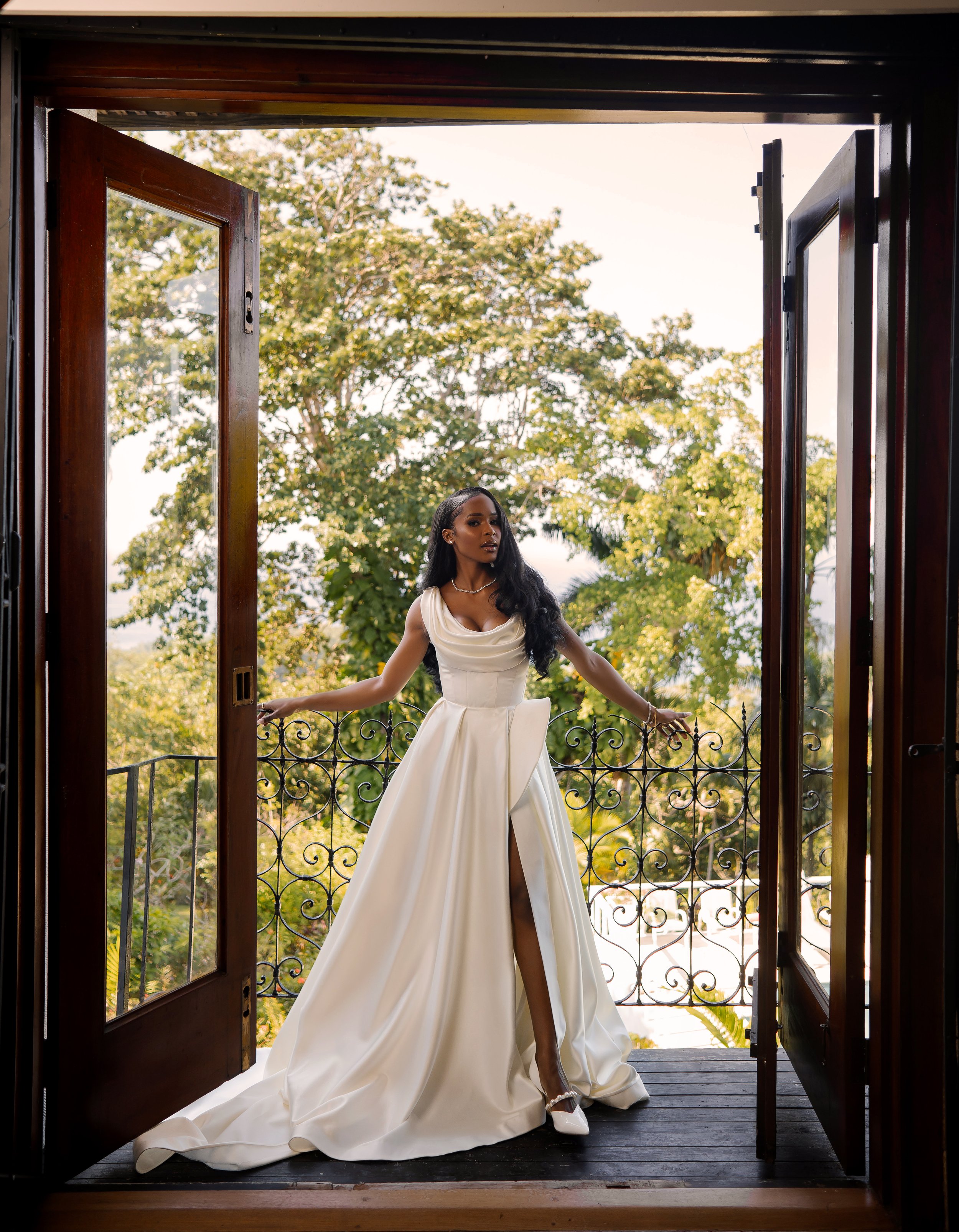 A woman in a white gown standing in a doorway, with trees and sky visible in the background.