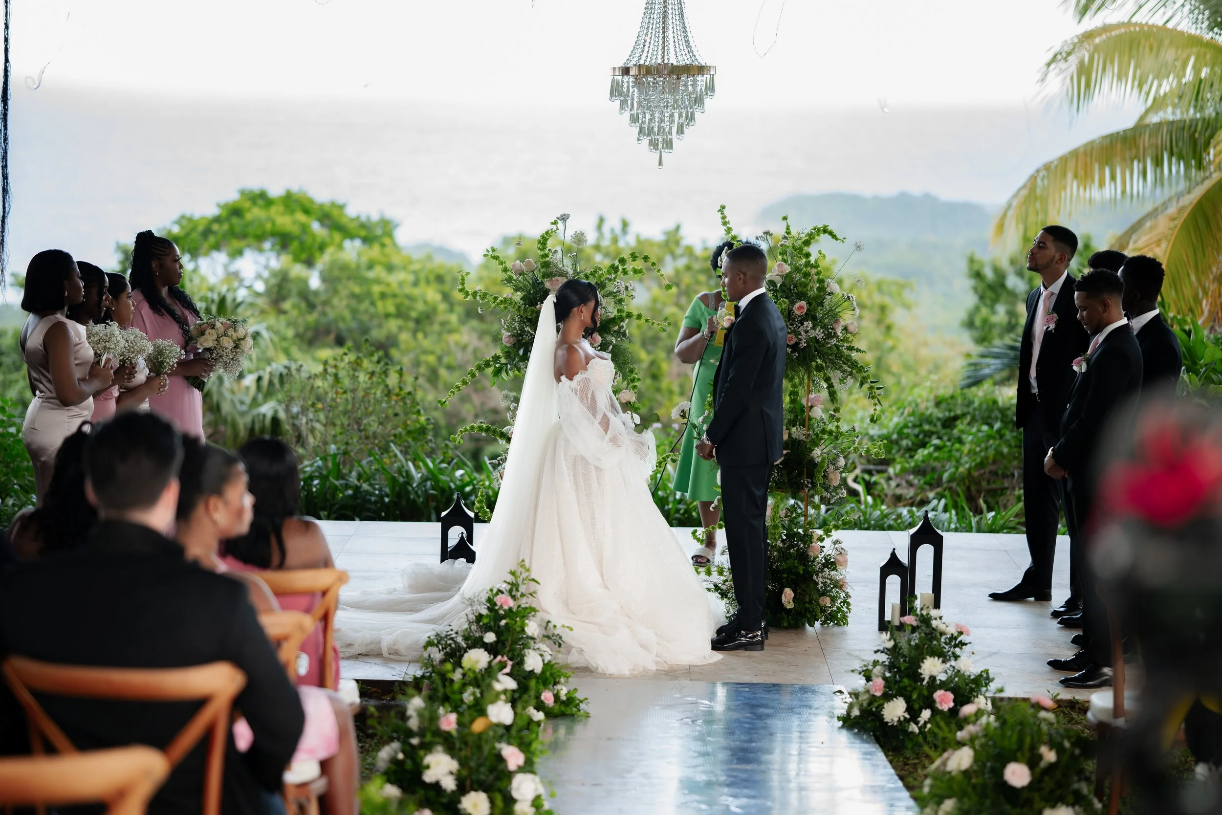 A wedding ceremony taking place outdoors with a bride and groom standing facing each other, surrounded by floral arrangements. Bridesmaids and groomsmen are standing on either side, with lush greenery and trees in the background and a chandelier hang