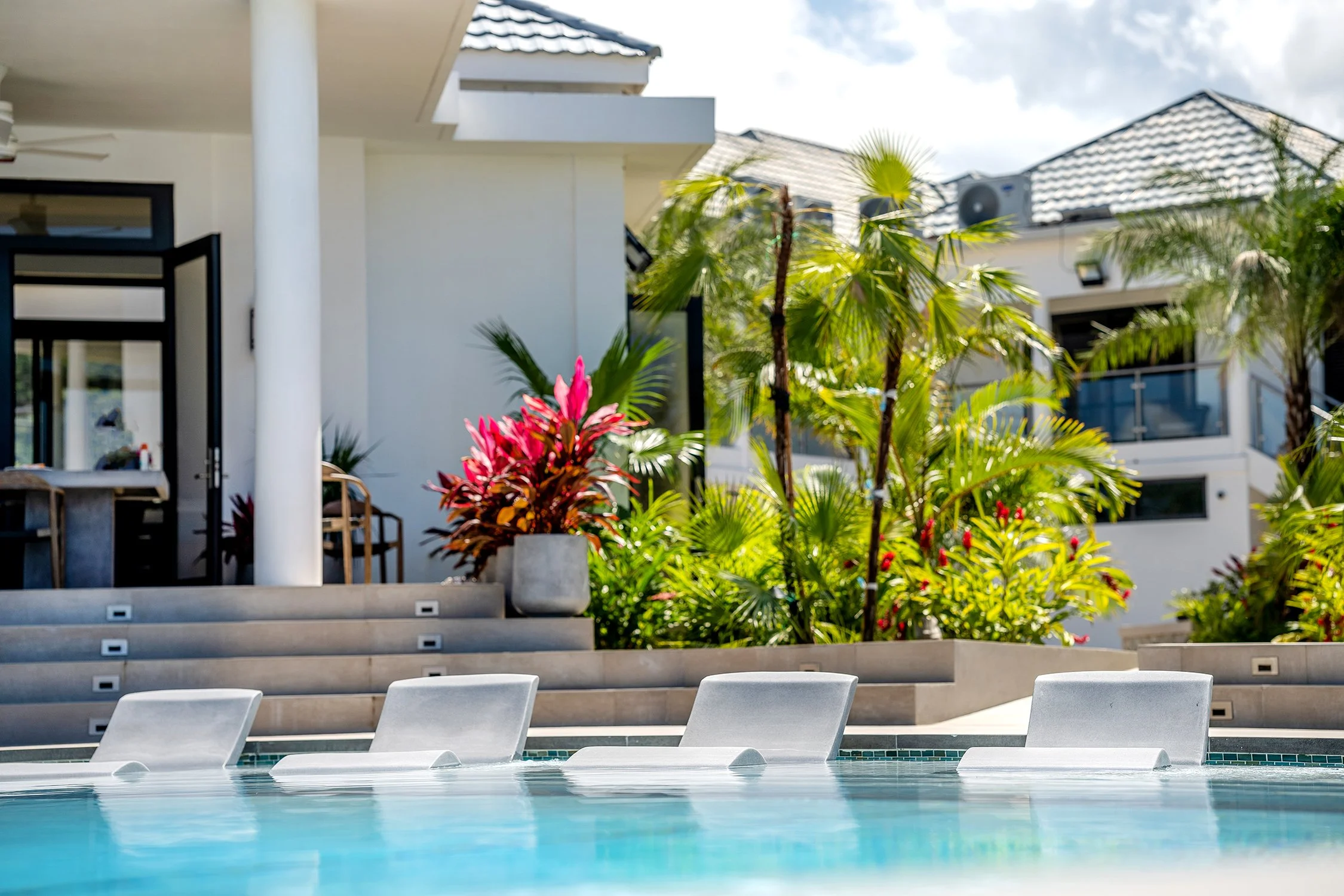 View of a modern swimming pool area with white lounge chairs, lush green palm plants, and residential buildings with tiled roofs in the background.