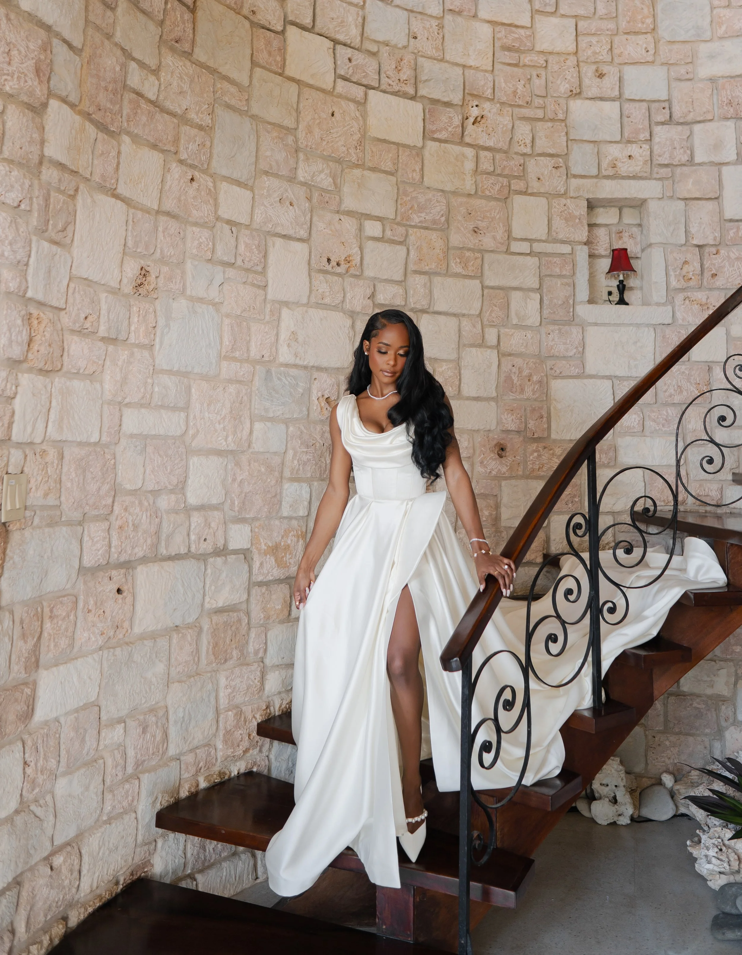 A woman in a white dress with a slit is sitting on a dark wood staircase with black wrought iron railing, against a beige stone wall, near a small recessed shelf with a red lamp.