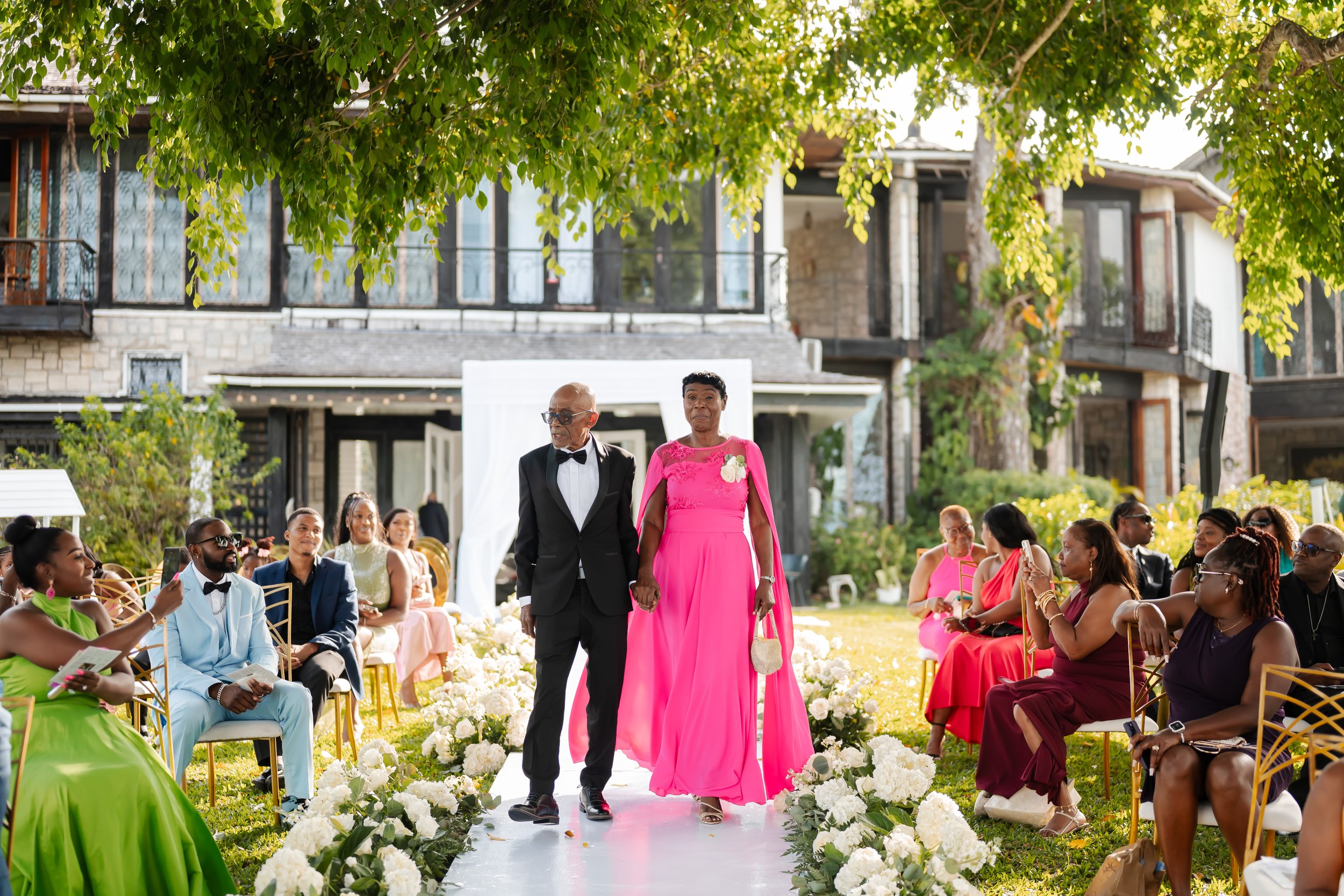An elderly man and woman walking down an outdoor wedding aisle. The man wears a black tuxedo, glasses, and a bow tie, while the woman is dressed in a bright pink gown, holding a small handbag. Guests are seated on either side of the aisle, which is d
