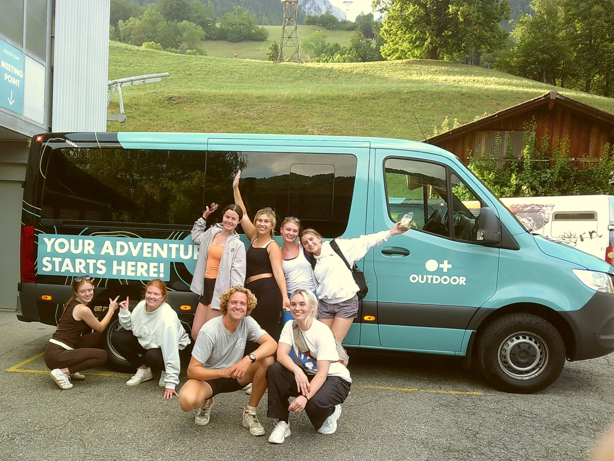 A group of young women and one man posing in front of a blue van with the text 'Your Adventure Starts Here!' on the side, in an outdoor parking lot with a scenic green hillside and trees in the background.