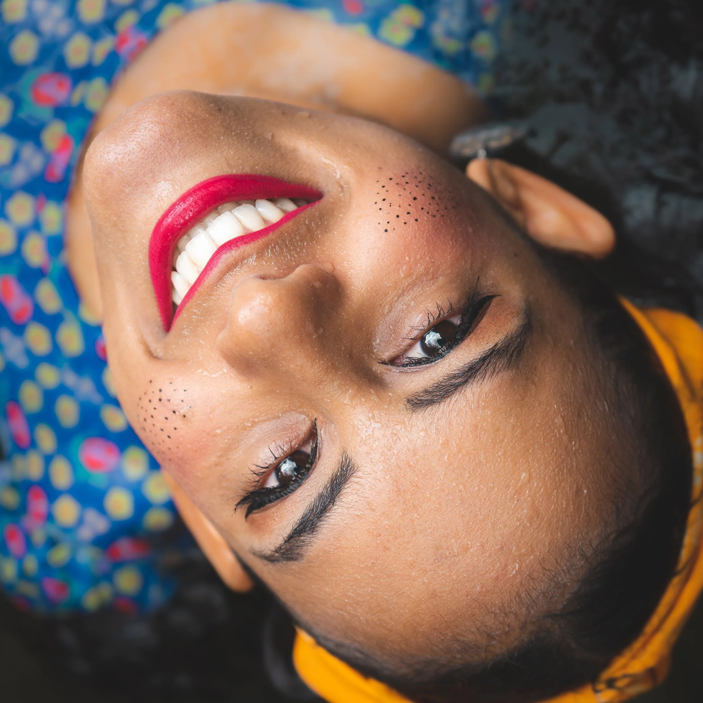 A woman with dark hair and brown skin lying down, smiling, with bright red lipstick, eye makeup, and small black freckles on her cheek, wearing a colorful patterned shirt and an orange headband.