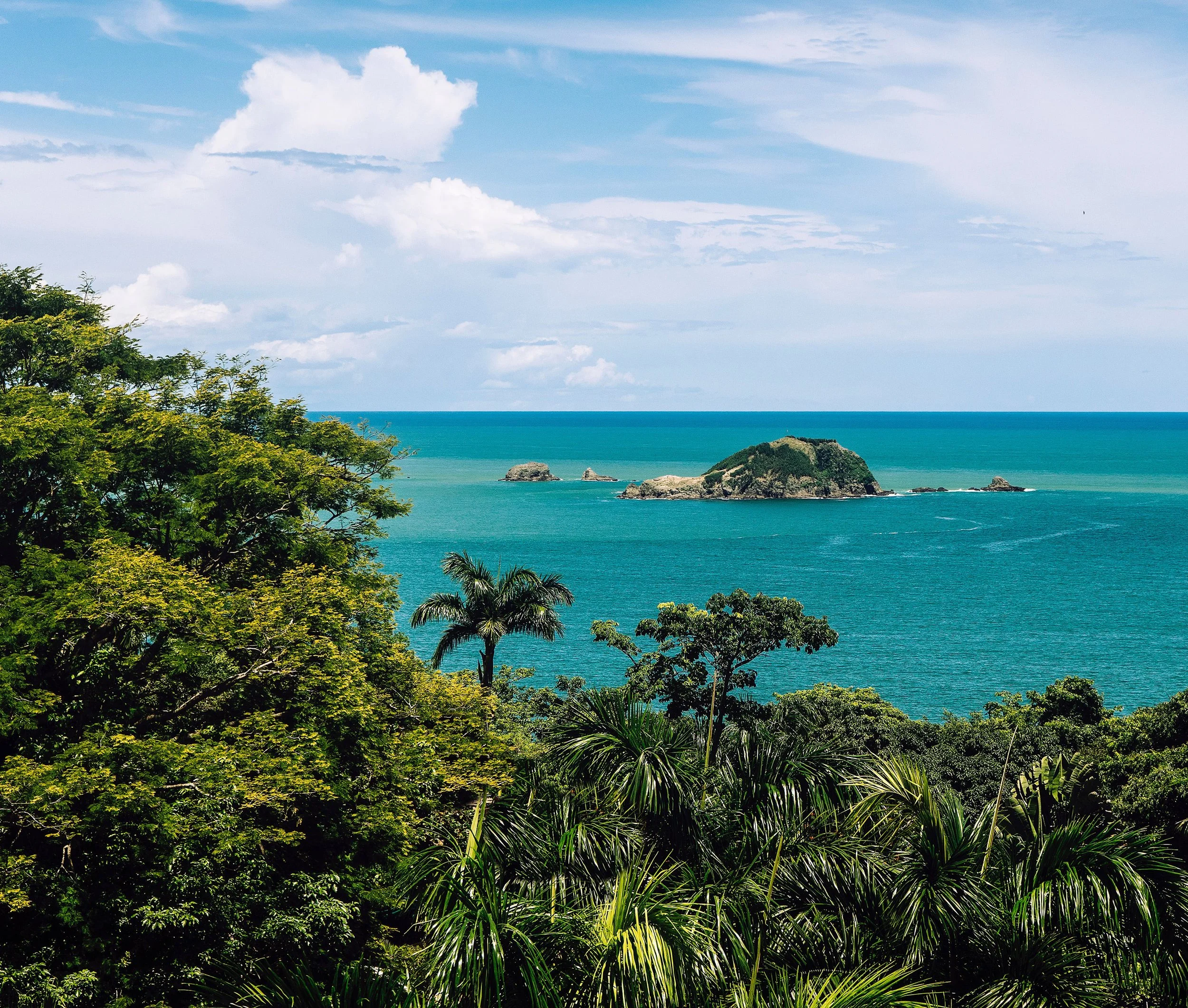 Tropical landscape with green trees in the foreground, a large ocean with small islands in the background, and a partly cloudy sky.