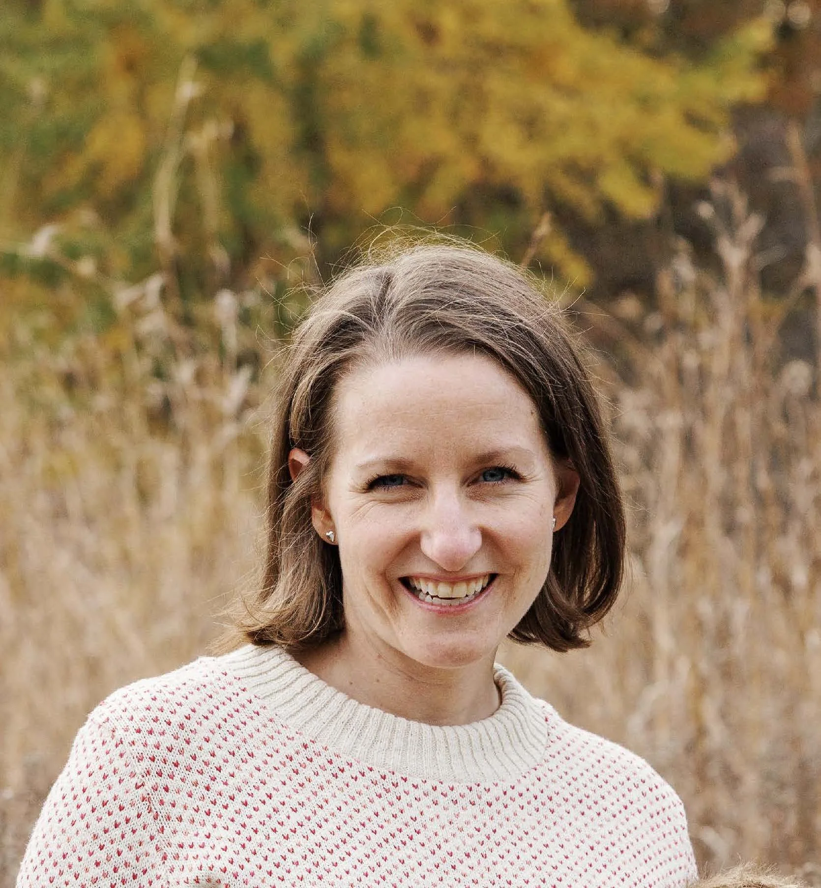 A woman with shoulder-length brown hair, smiling, standing outdoors with a blurred background of yellow and green foliage, wearing a cream sweater with red hearts.