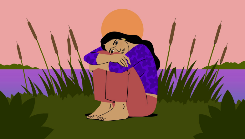 A woman sitting on grass near a lake at sunset, with arms wrapped around her knees and a sad expression.