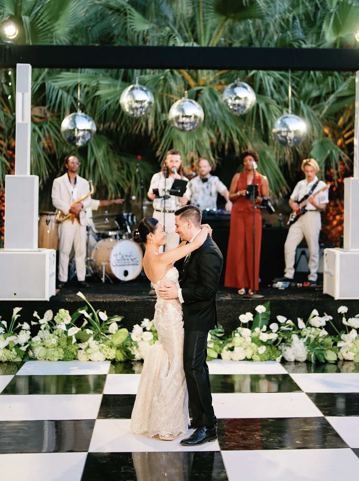 Bride and groom on black and white checkered dance floor with band in the background underneath disco ball stage 