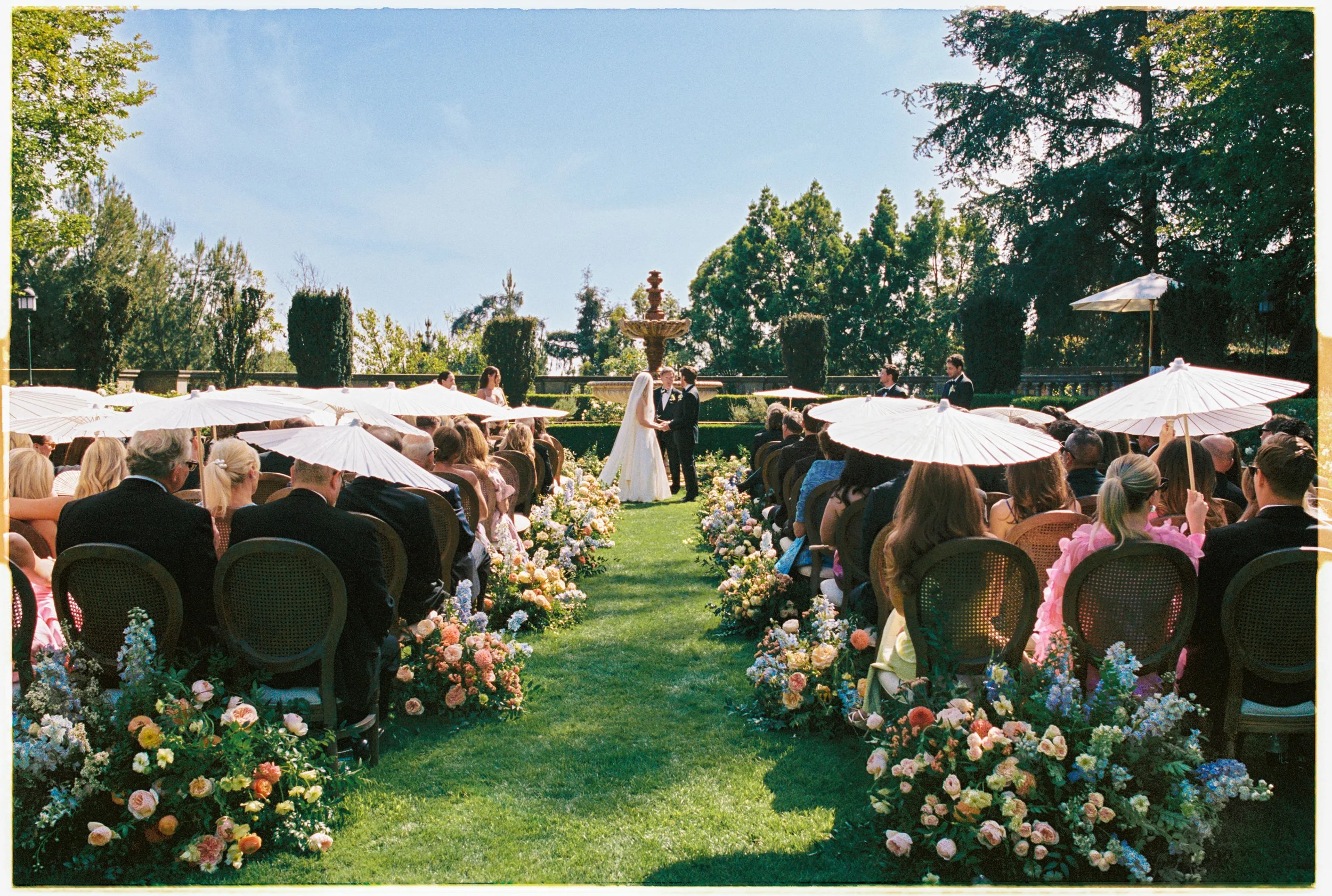 Ceremony with guests holding white parasols and flowers on the grass 