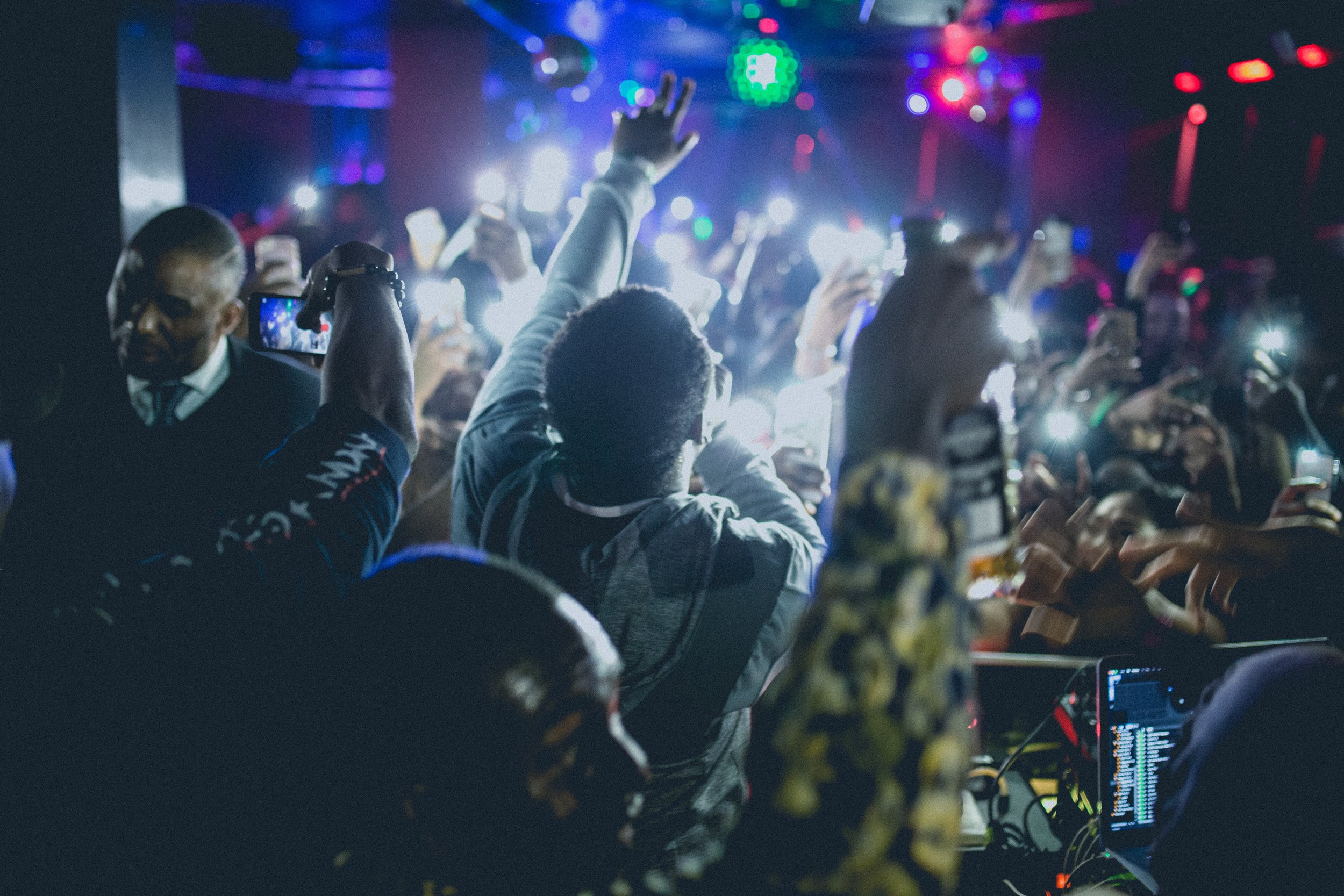 Menschen auf einer Party tanzen und machen Fotos mit Smartphones, bunte Lichter und DJ-Equipment im Hintergrund.