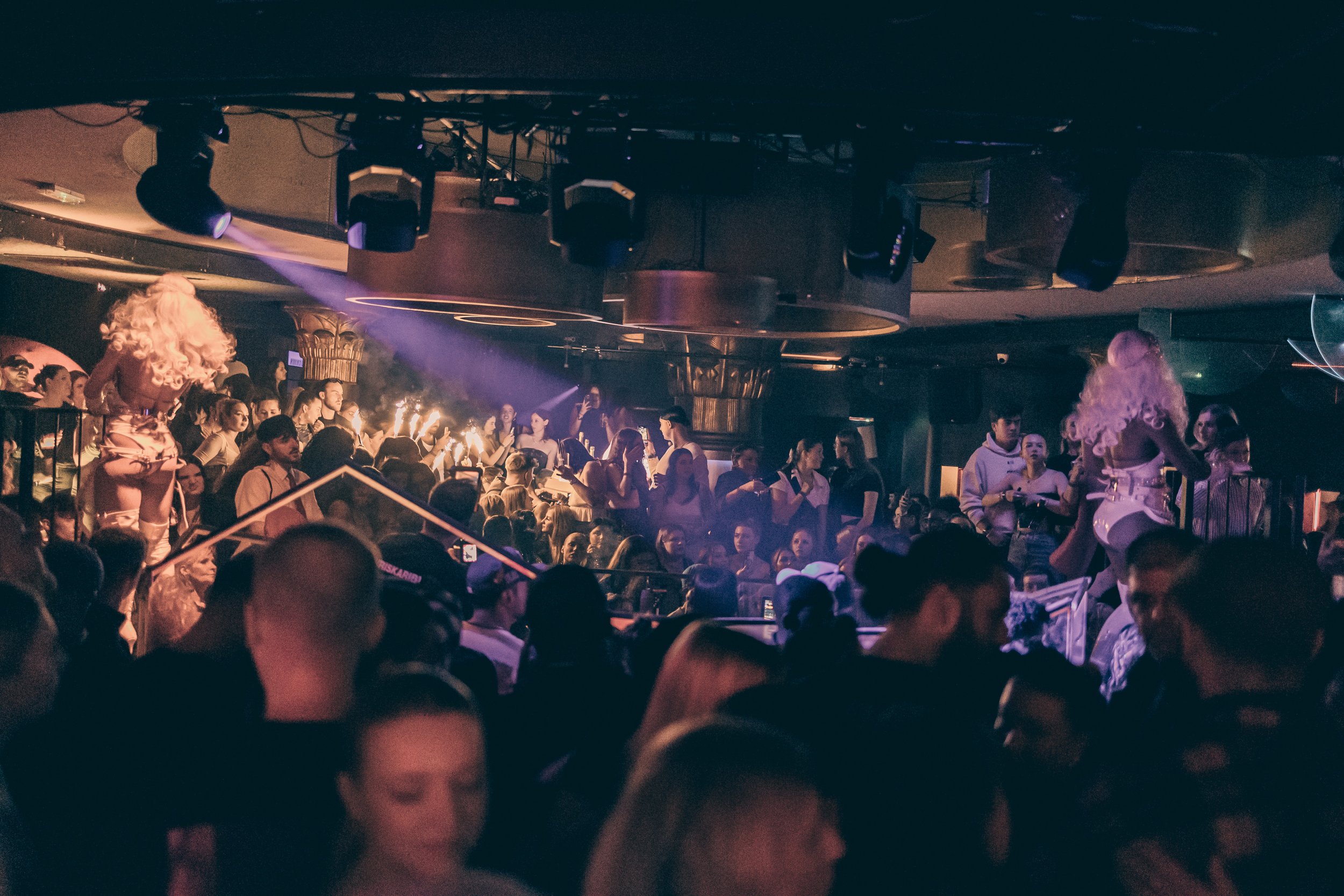 Menschen in einer Diskothek bei einer Party, mit einem DJ-Pult und Tanzflächen, das Licht ist dunkel mit bunten Lichteffekten.