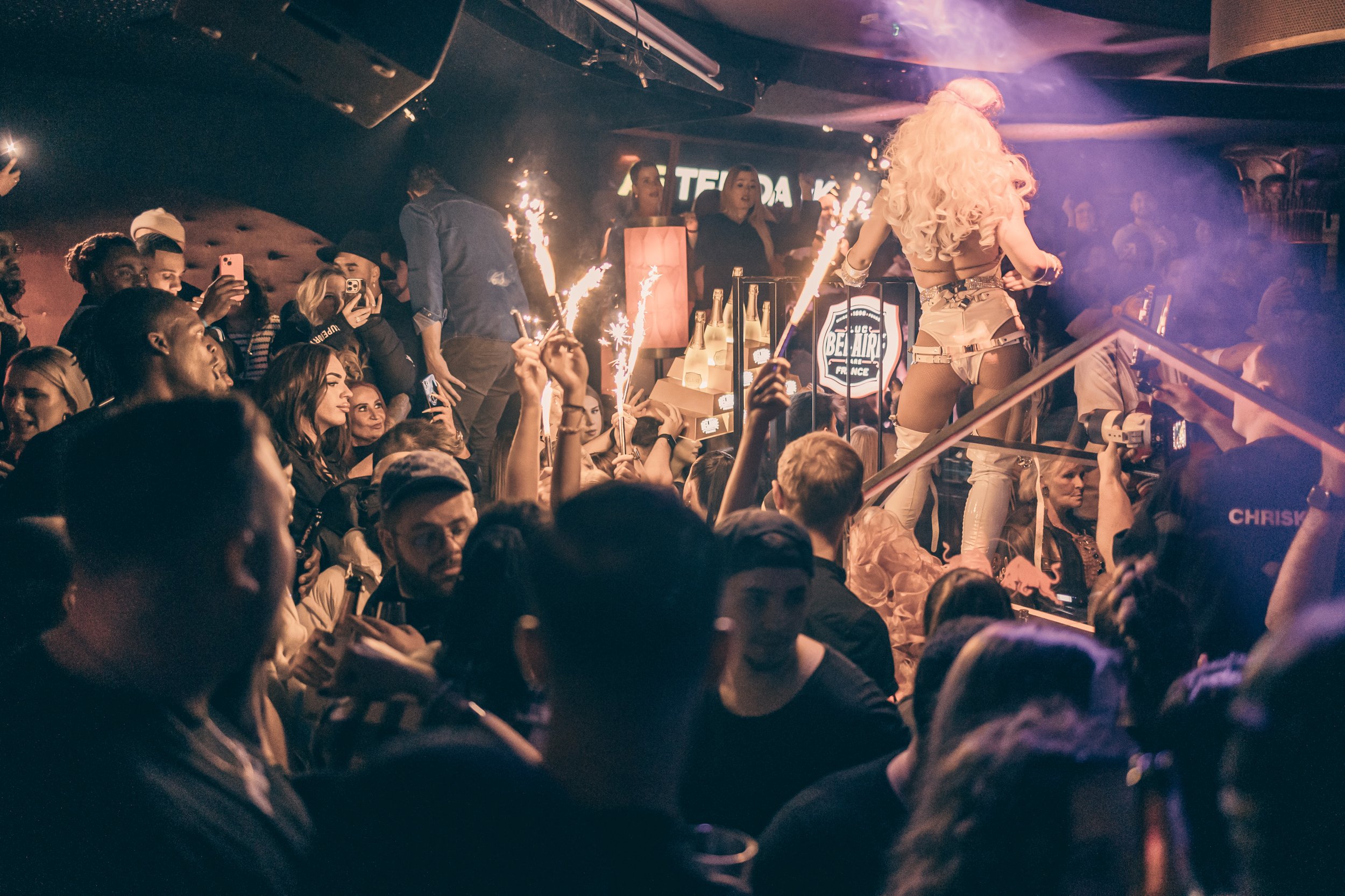 Menschen in einer angesagten Club- oder Partykneipe feiern mit einer Frau auf einer Bühne, die mit langen blonden Haaren und in auffälliger Kleidung posiert. Es werden Kerzen und Lichterströme verwendet, mehrere Personen nehmen Fotos oder Videos auf.