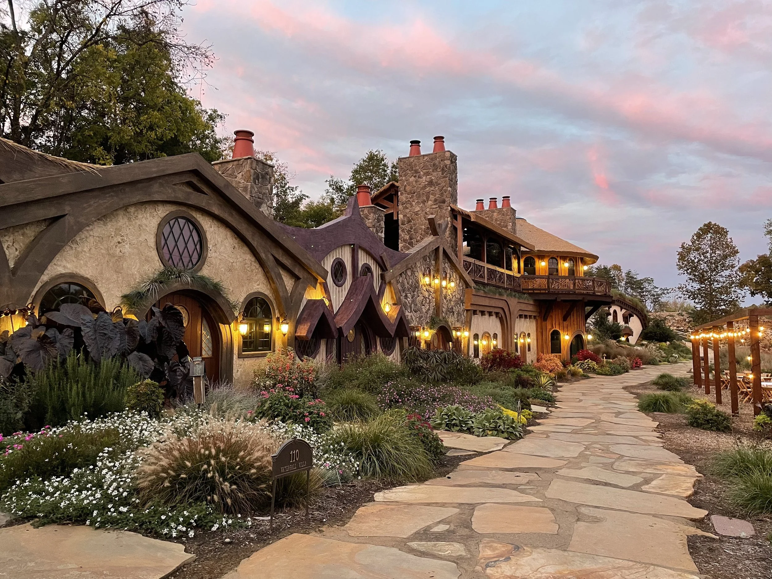 A picturesque, fantasy-style house with multiple stone chimneys, wooden balconies, and curved, mushroom-like roof sections illuminated by exterior lights at dusk, surrounded by a landscaped garden with flowering bushes and a stone-paved pathway.