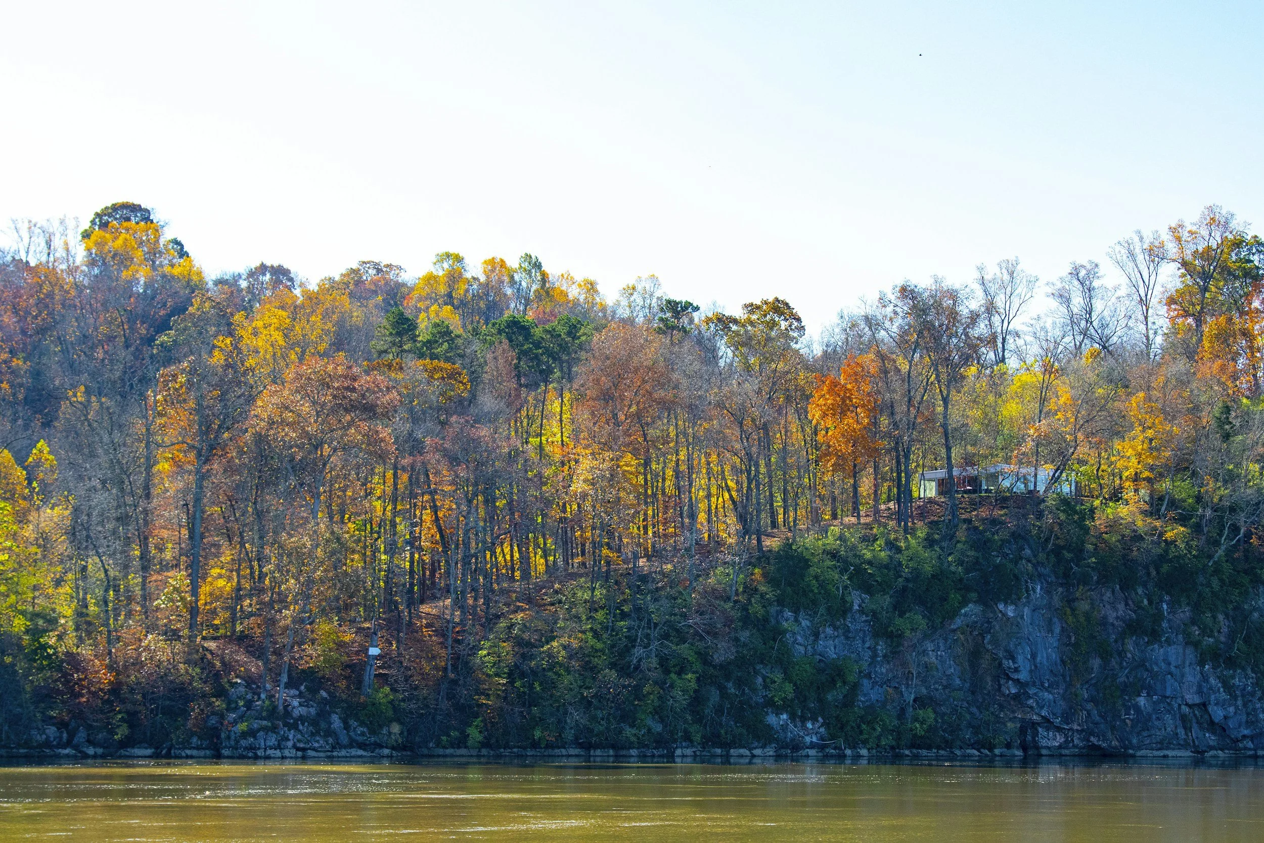 Autumn landscape with trees in fall colors along a riverbank, with a house partially visible among the trees.