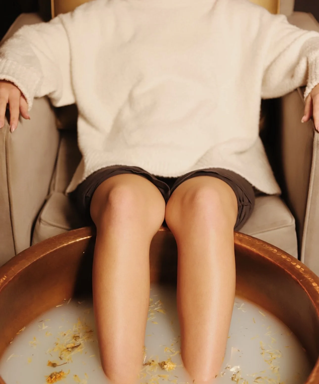 Person sitting in a chair with feet submerged in a copper tub filled with milky water and flower petals.