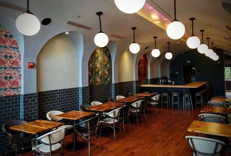 Empty restaurant with wooden tables, black and white chairs, black tiled walls, arched accents, colorful wallpaper, and spherical hanging lights.