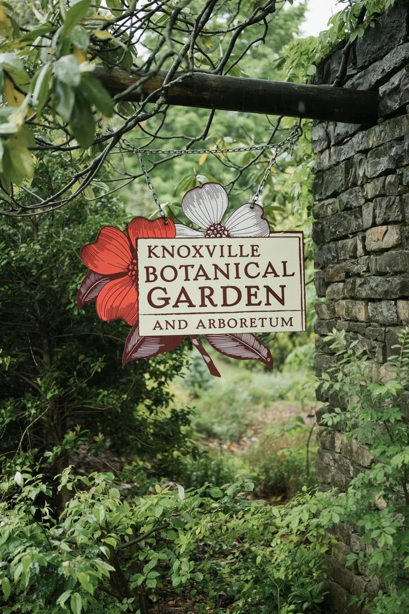 Sign for the Knoxville Botanical Garden and Arboretum hanging among green plants and trees, with a stone wall on the right.