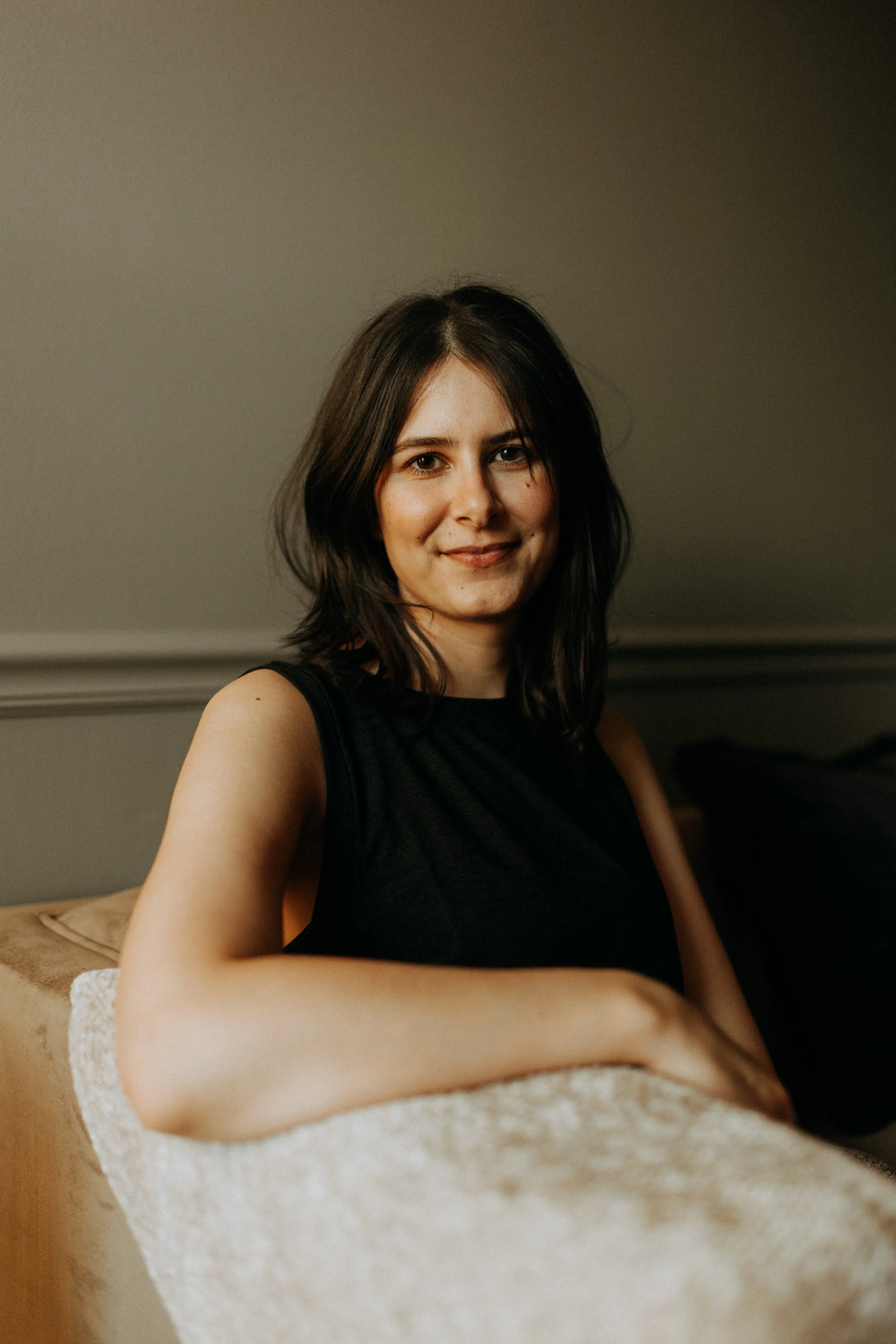 A young woman with shoulder-length dark hair, wearing a black sleeveless top, sitting on a beige sofa, smiling at the camera in a dimly lit room.
