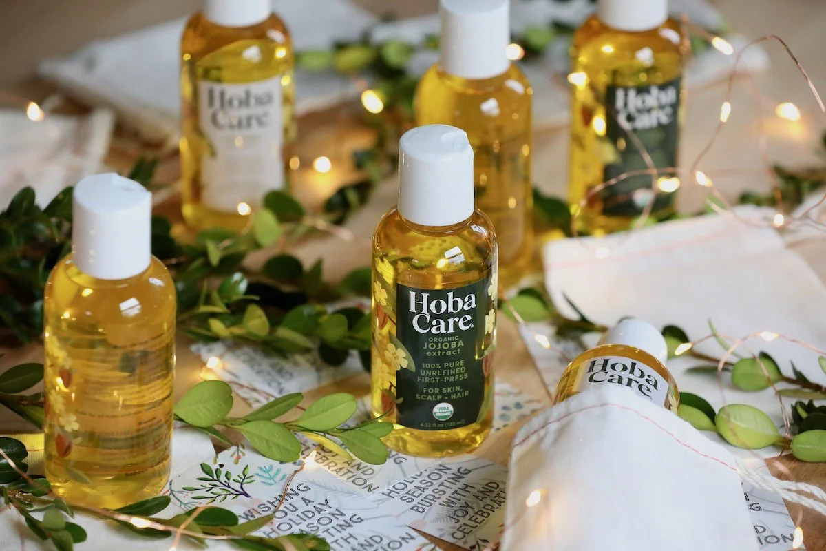 Six sample-sized bottles of jojoba oil arranged artfully on a surface. The scene is scattered with LED lights and greenery.