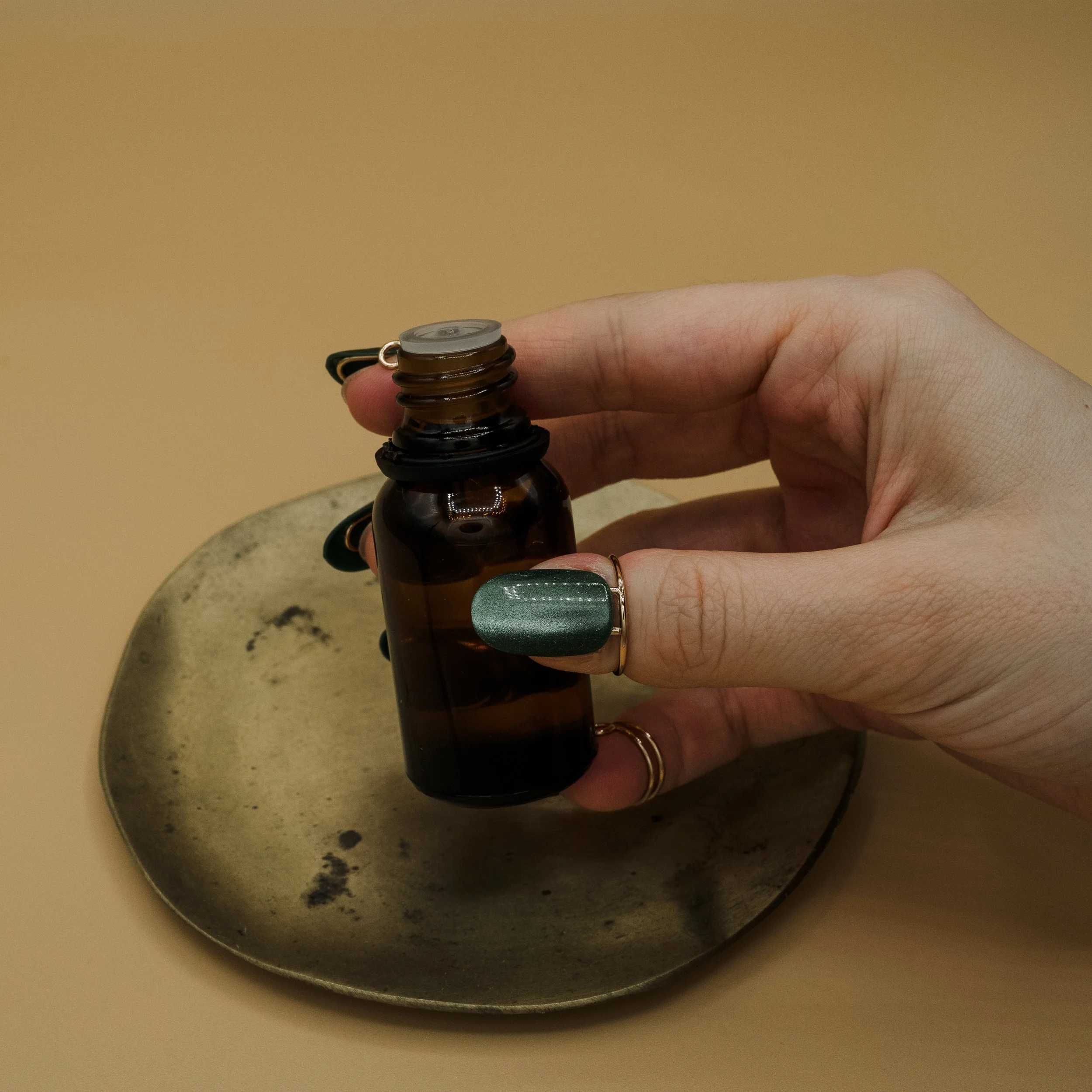 A hand holding an amber glass bottle with a gray cap, positioned above a small round metallic dish.