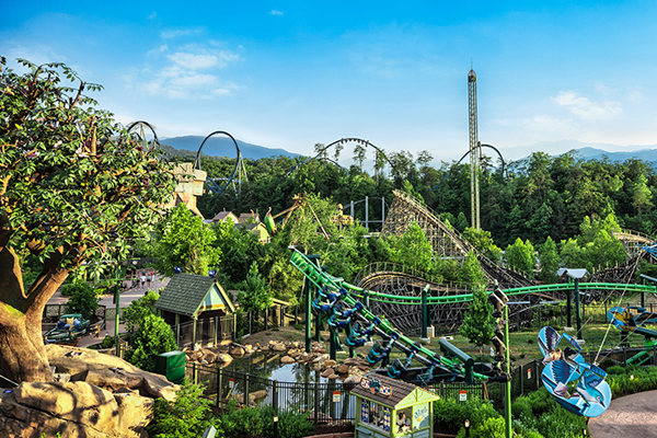 A view of an amusement park with roller coasters and trees under a blue sky.