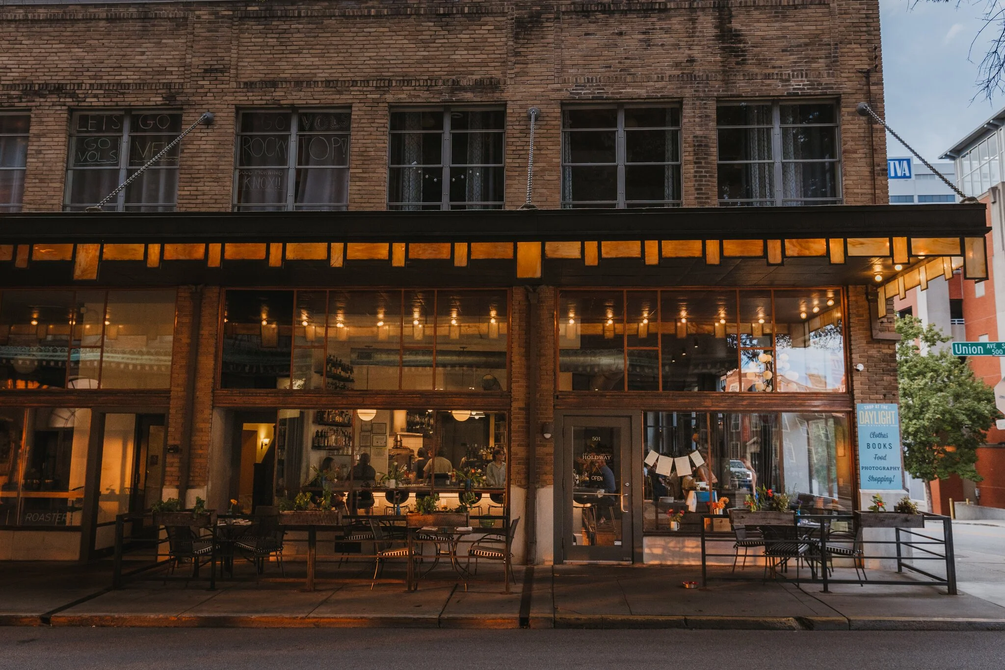 A two-story brick building with large windows on both floors, housing a warmly lit restaurant or cafe with outdoor tables and chairs on the sidewalk, and a street sign indicating the intersection of Union Ave S and another street.