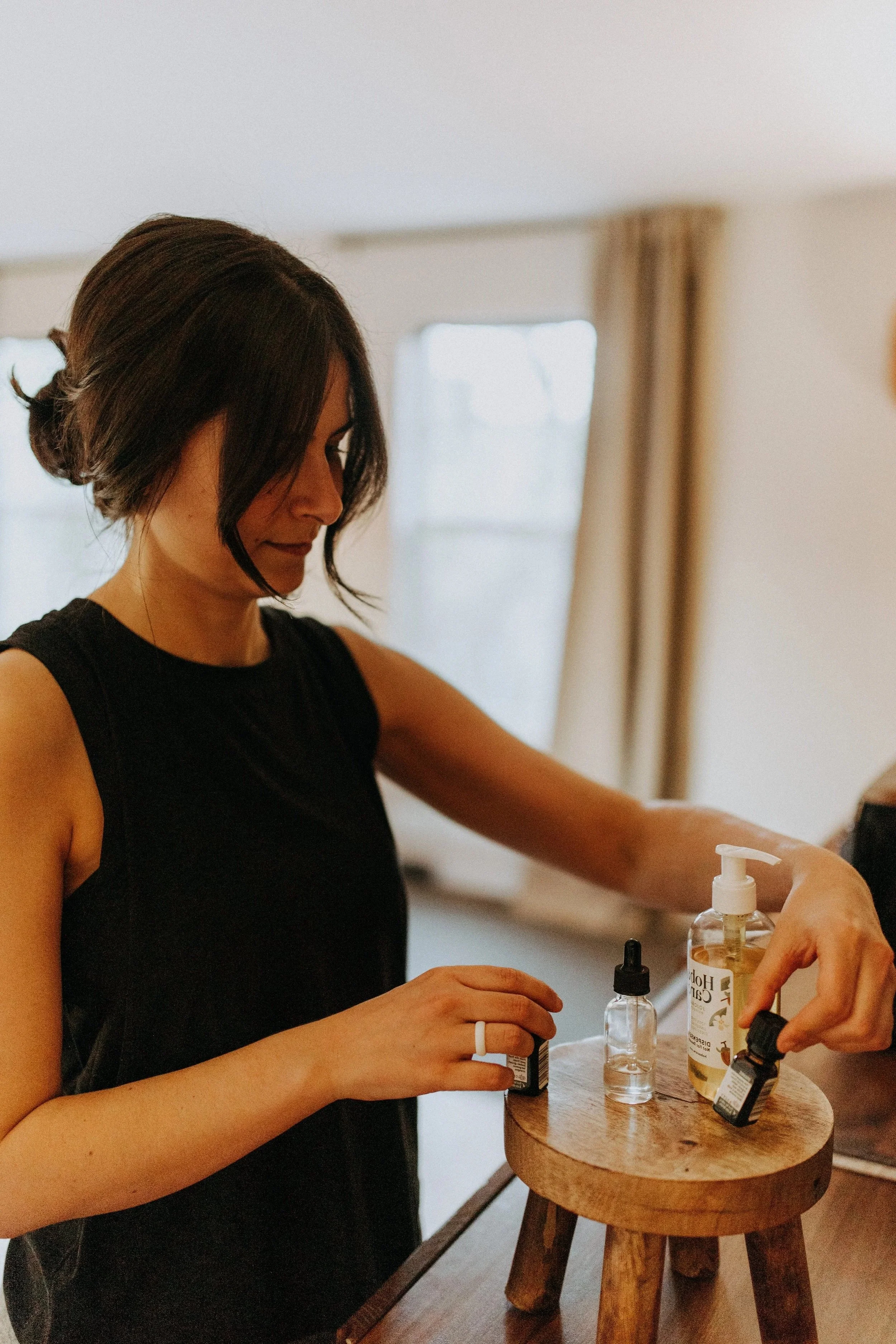 A woman with short dark hair wearing a black sleeveless top is sitting at a wooden table, using various bottles of skincare or beauty products, including a pump bottle and dropper bottles.