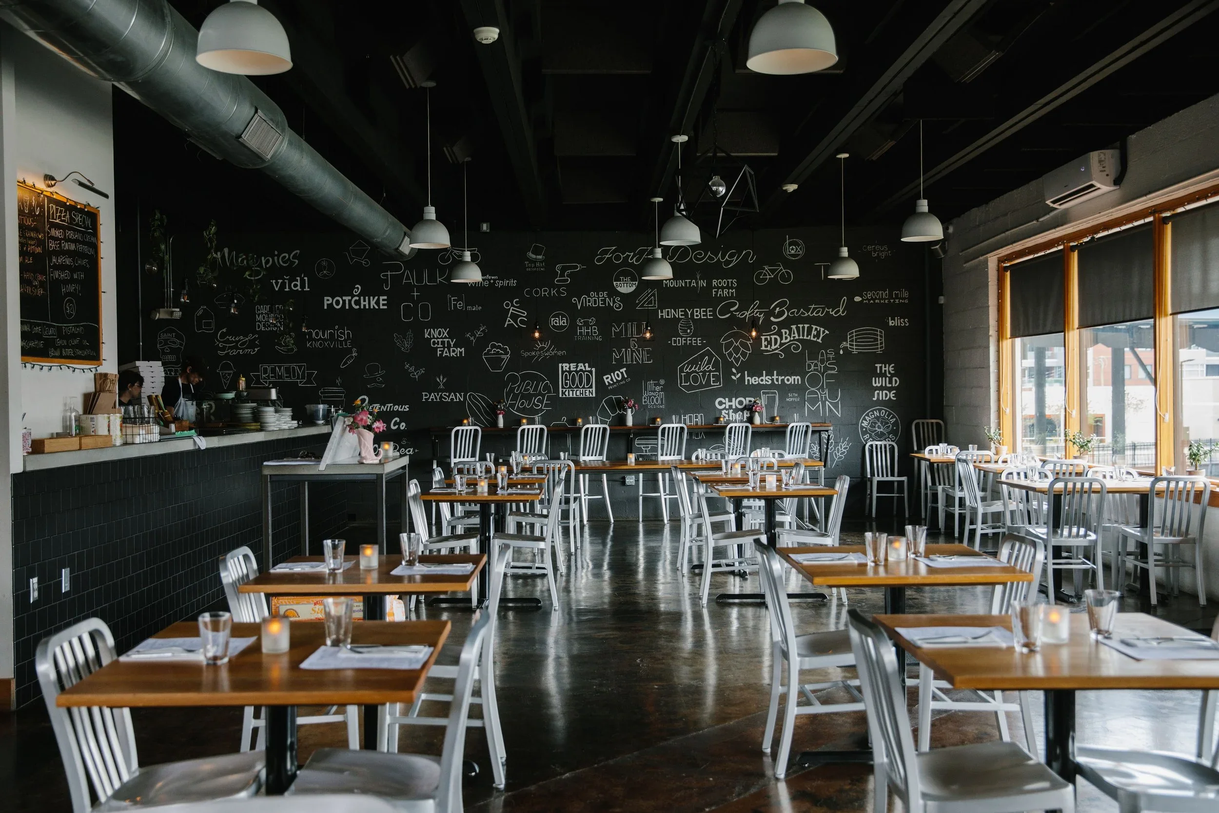 Empty restaurant with wooden tables, white chairs, a black chalkboard wall with menu drawings, and large windows with blinds.