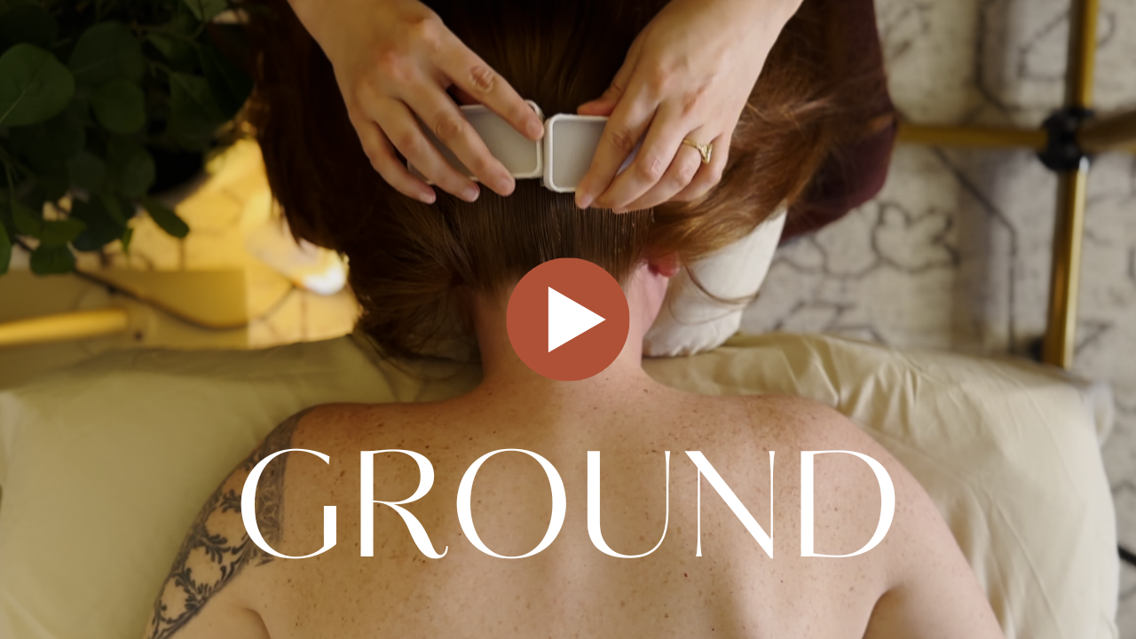 A person lying face down on a massage table, receiving a head massage, with a therapist pressing on their head. The setting appears to be a spa or massage therapy room, with a plant and a lamp visible in the background. The word "GROUND" is overlaid on the bottom of the image.