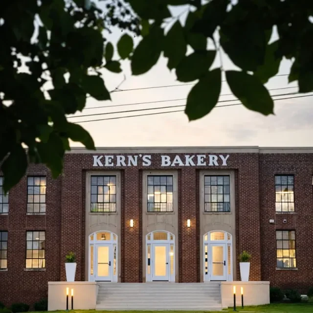 Brick bakery building with the sign 'KERN'S BAKERY' on top, three arched doors, stairs leading up to the entrances, and lit outdoor lamps, framed by tree branches and leaves at the top.