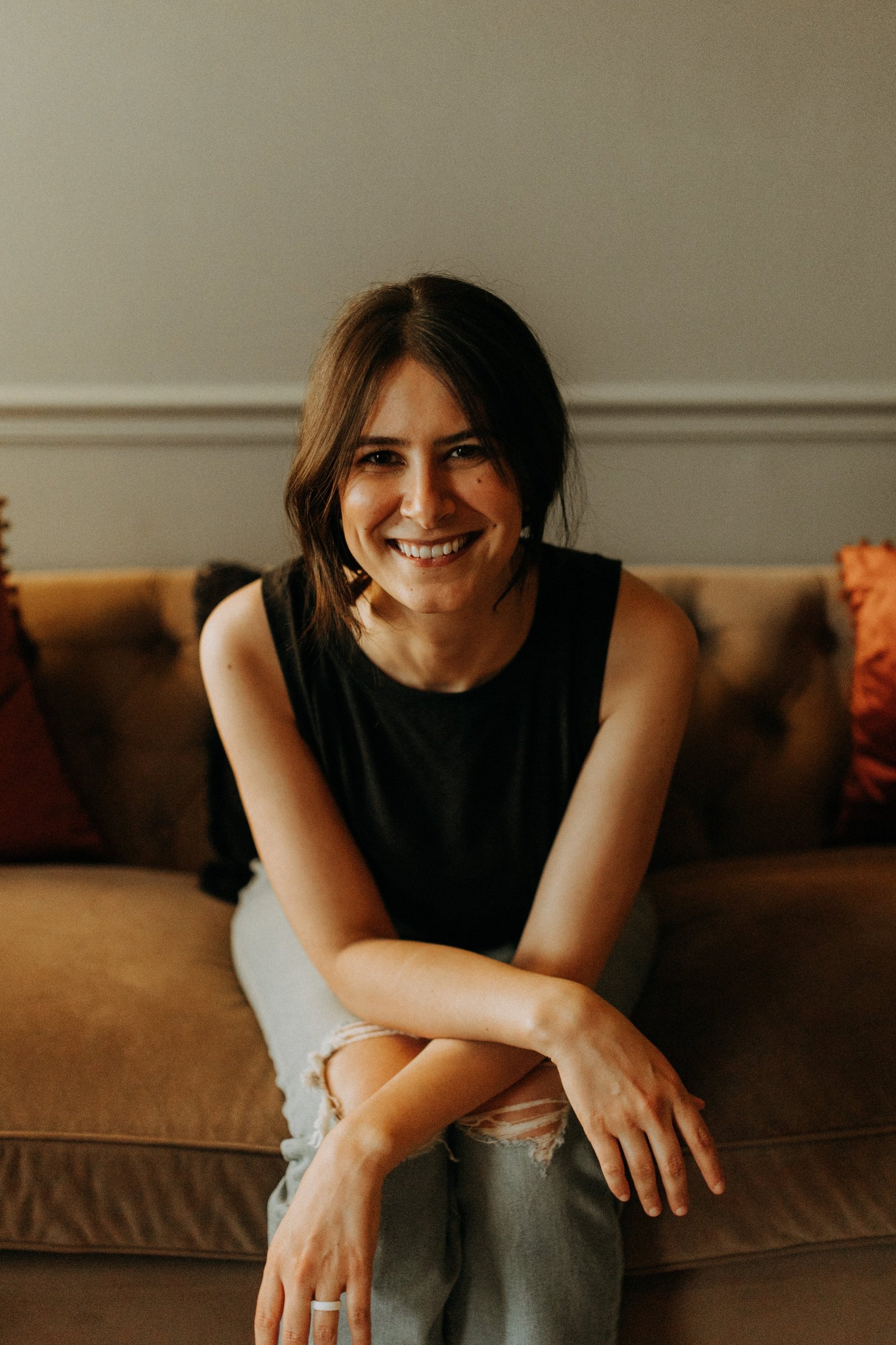A woman with dark hair and a black sleeveless top sitting on a brown couch, smiling at the camera.