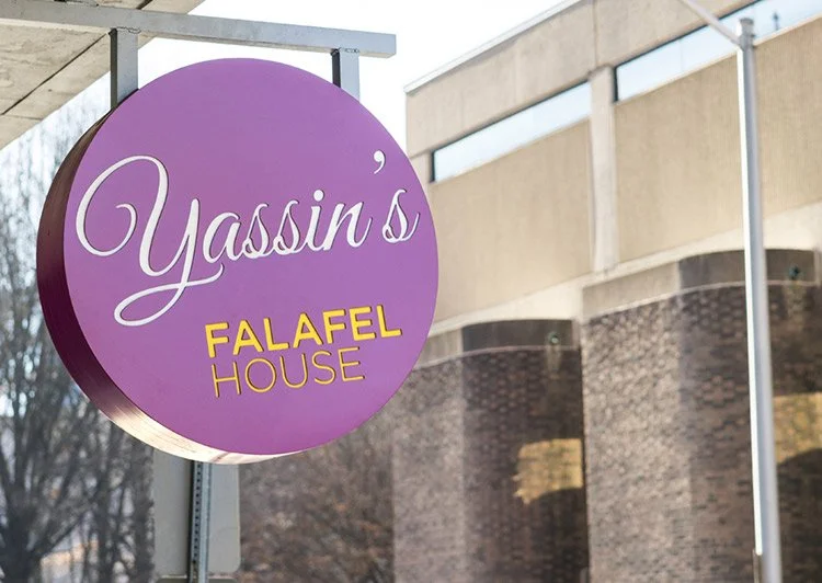 Close-up of a purple circular sign for Yassin's Falafel House with yellow text, mounted on a metal pole, with a building and trees in the background.