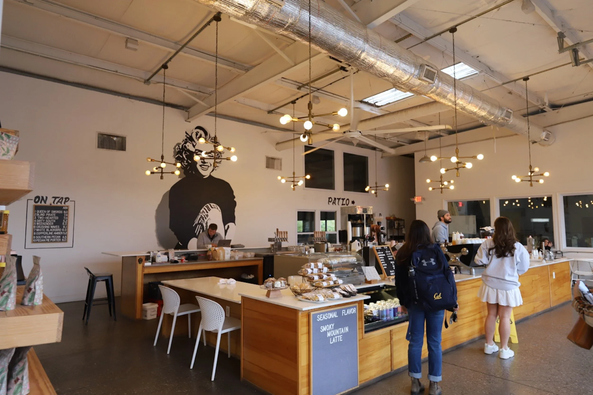 Interior of a modern coffee shop with a large black and white mural of a smiling woman on the wall behind the counter. Customers are ordering at the counter, which displays baked goods and a sign advertising seasonal latte flavors. The ceiling has exposed ductwork and hanging light fixtures.