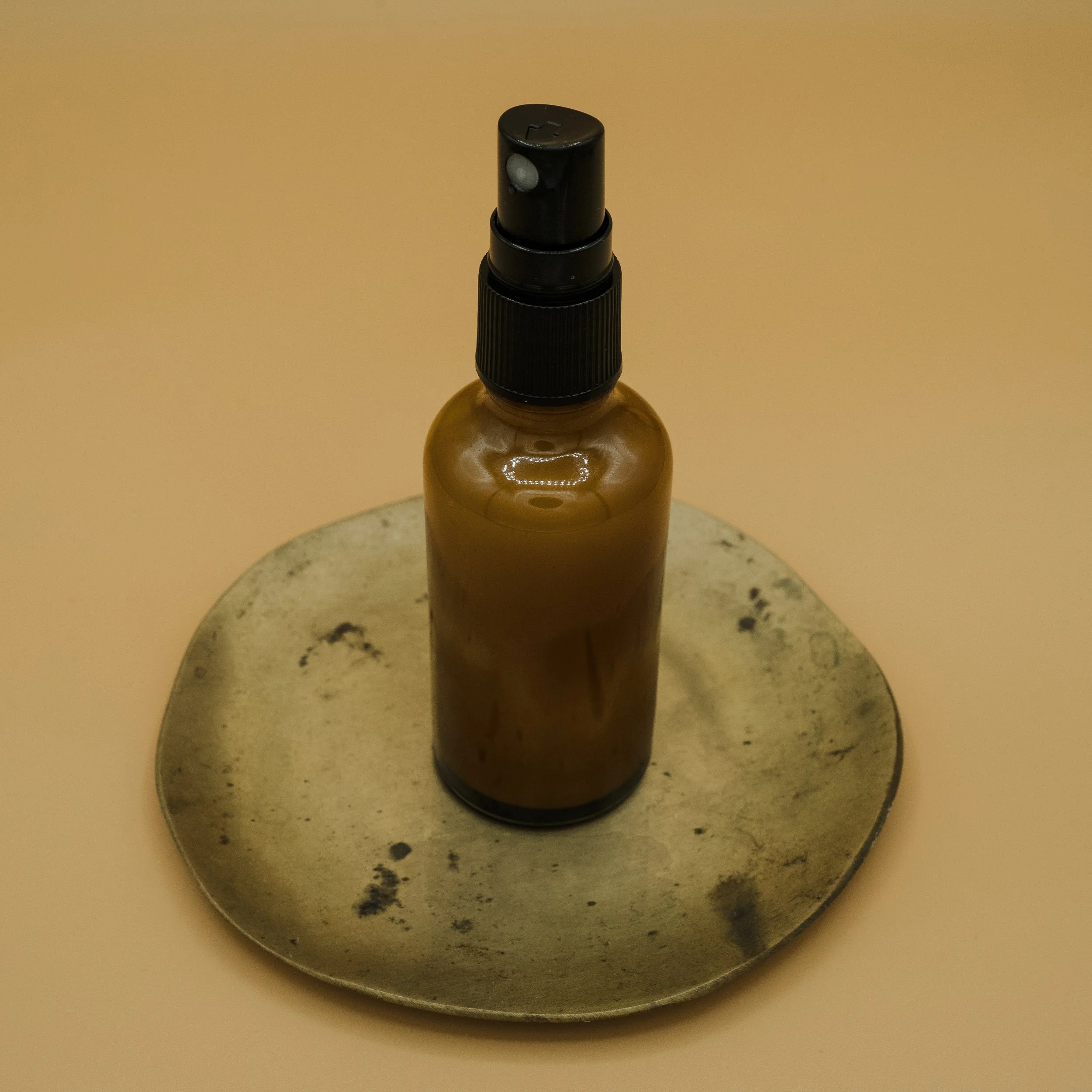 Brown spray bottle with a black spray nozzle on a reflective metallic tray on a beige surface.