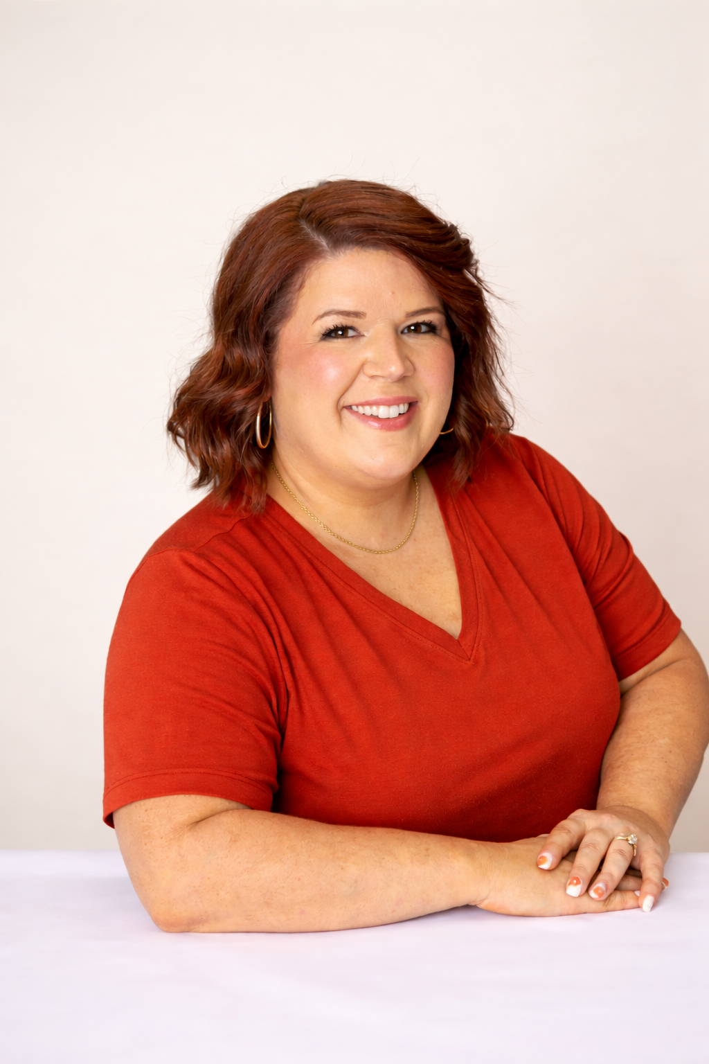 A woman with shoulder-length red hair, wearing a red V-neck shirt, gold hoop earrings, a gold necklace, and a wedding ring, smiling at the camera against a plain light background.