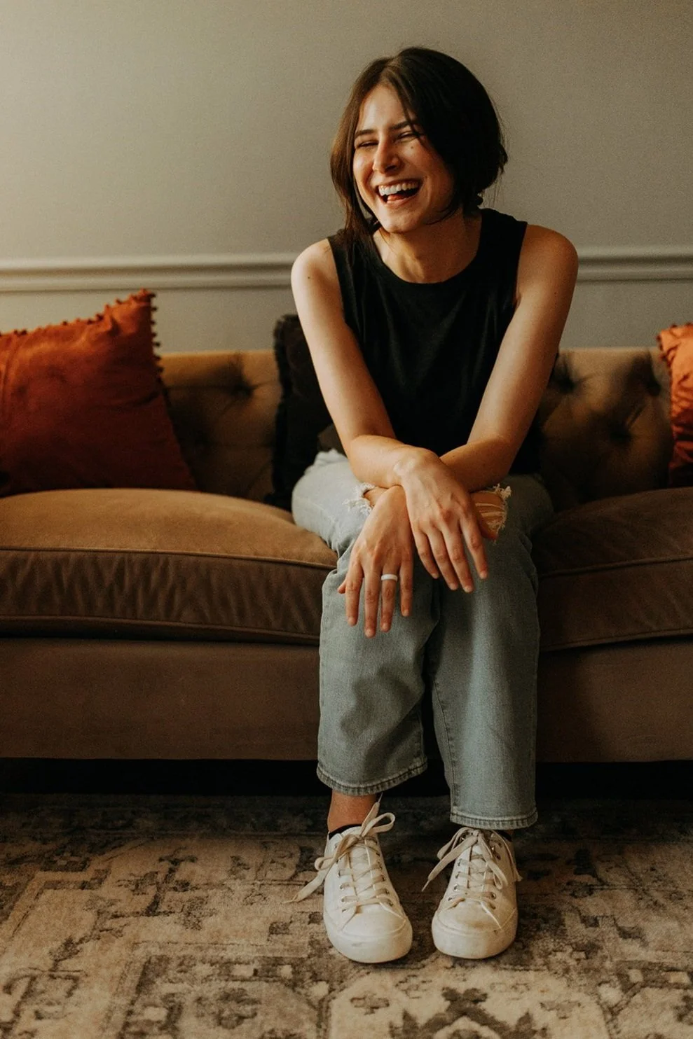 A young woman with short dark hair, wearing a black sleeveless top, ripped jeans, and white sneakers, sitting on a brown couch, laughing with her eyes closed, in a cozy living room with orange and black cushions.