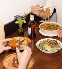 People enjoying a meal with two bowls of soup, salads, a beer, and a fried chicken drumstick on a plate