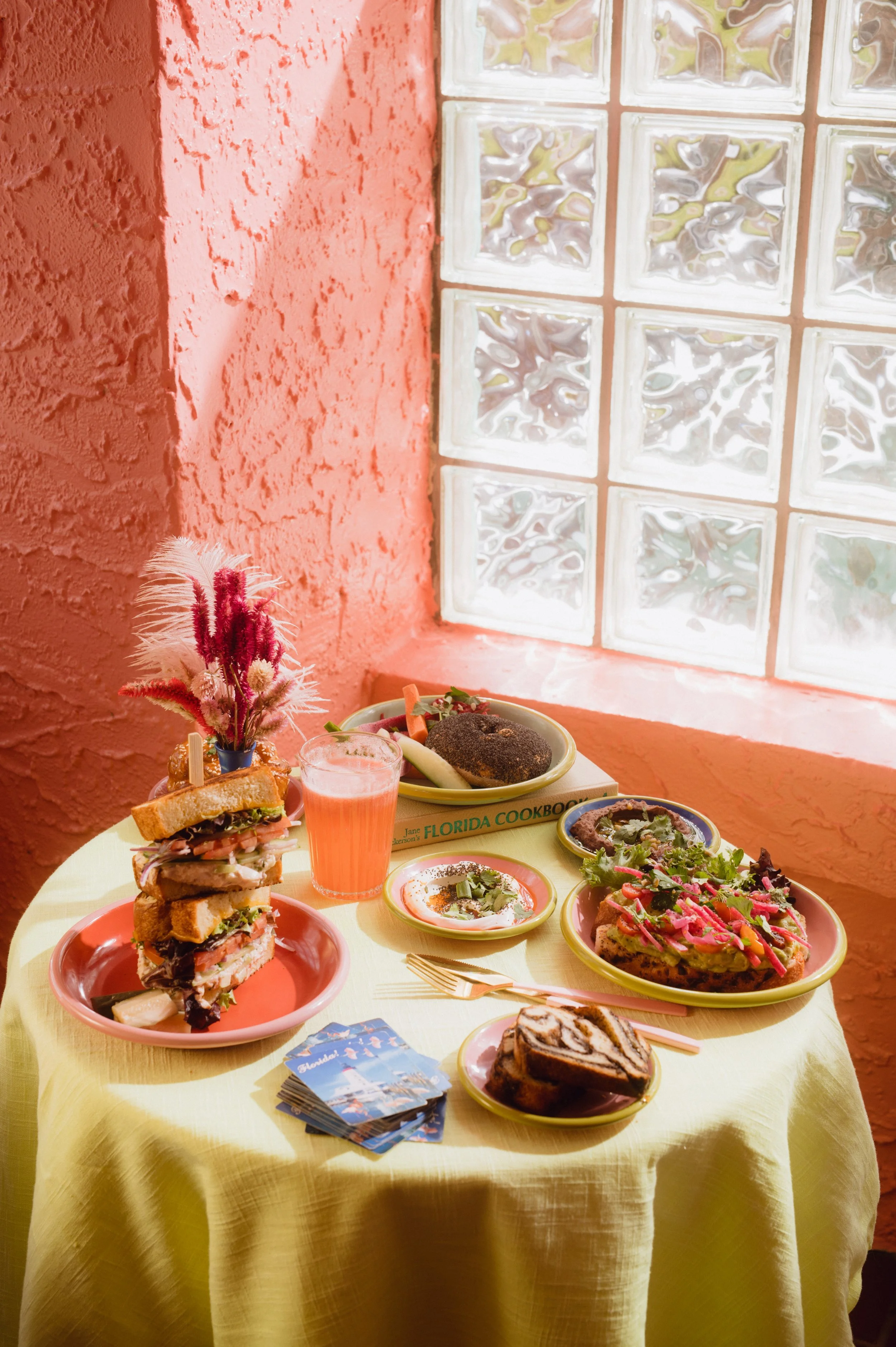 A table with various foods including a large stacked sandwich, a glass of pink beverage, a plate with fried food, a salad, a slice of cake, and a small dish with dip, set against a pink wall and a glass block window.