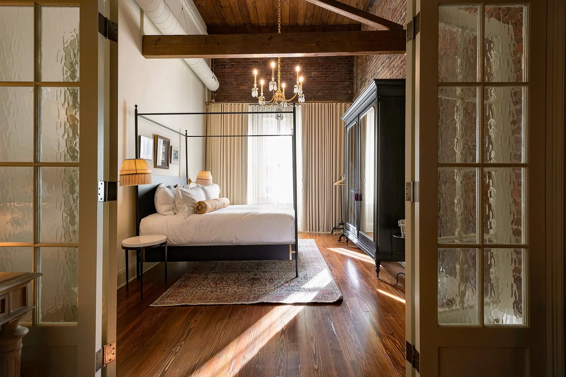 A bedroom with a four-poster bed, two bedside lamps, a black wardrobe, and a chandelier, with sunlight streaming through white curtains.