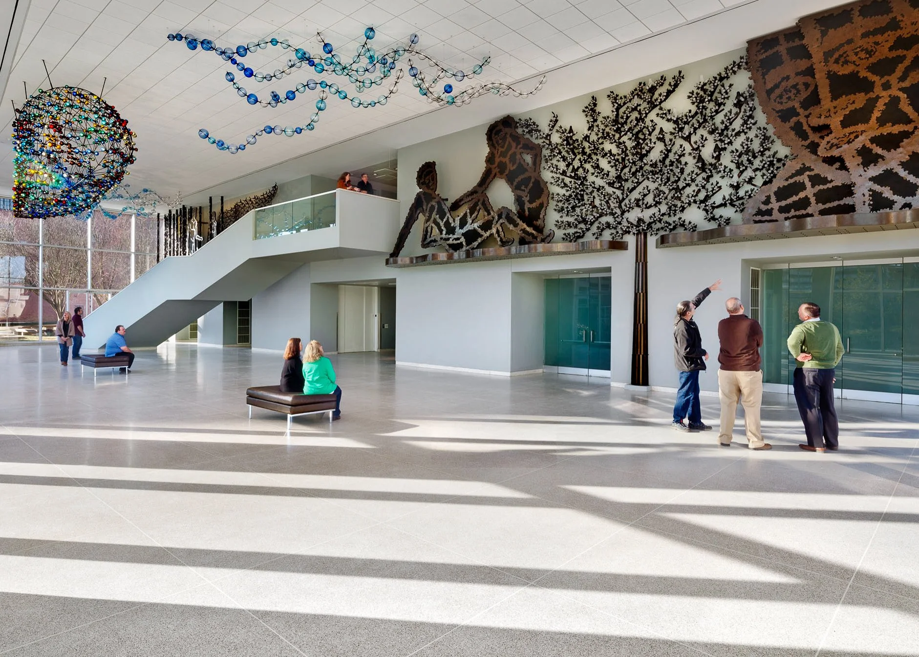 Interior of a spacious modern building lobby with high ceilings, large windows, and artwork on the walls. Several people are seated and standing, engaged in conversation, with sunlight casting shadows on the floor.