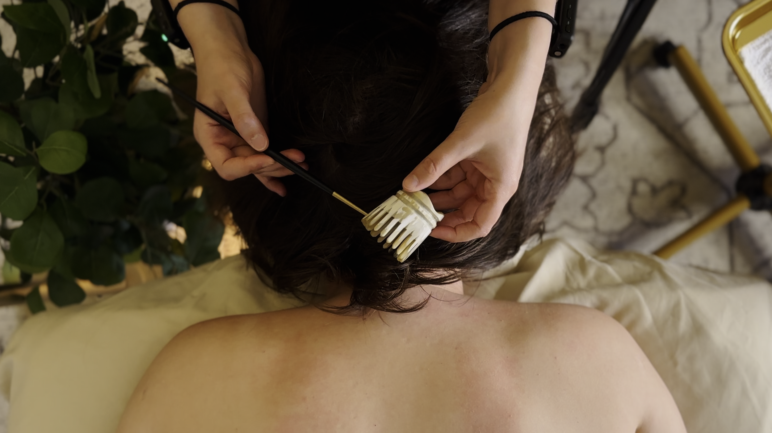 An overhead view of a client lying face down on the massage table. A massage therapist holds a chopstick and horn scalp massager over the head.
