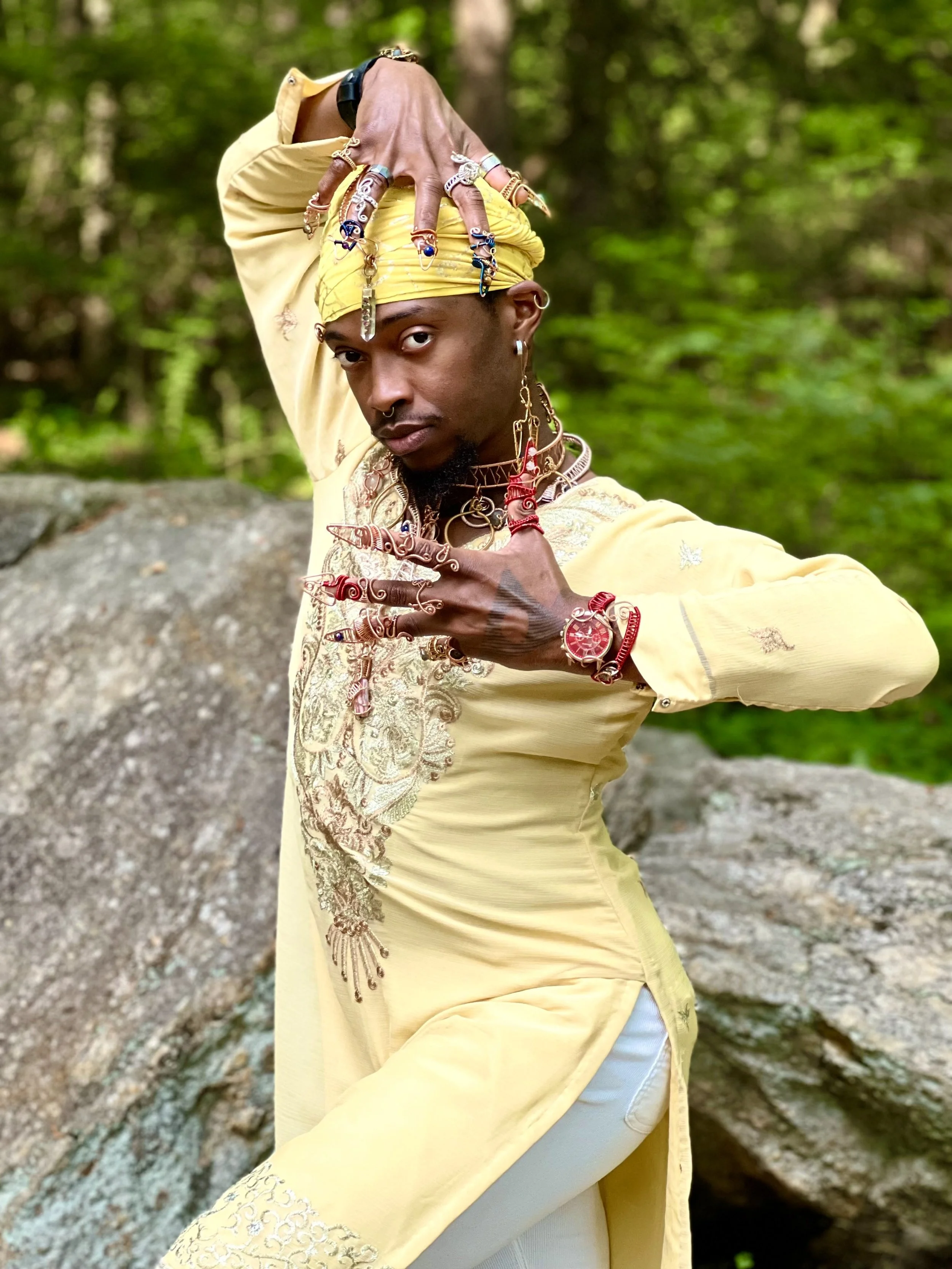 A person dressed in a yellow outfit with jewelry and accessories, posing outdoors with a forest background.