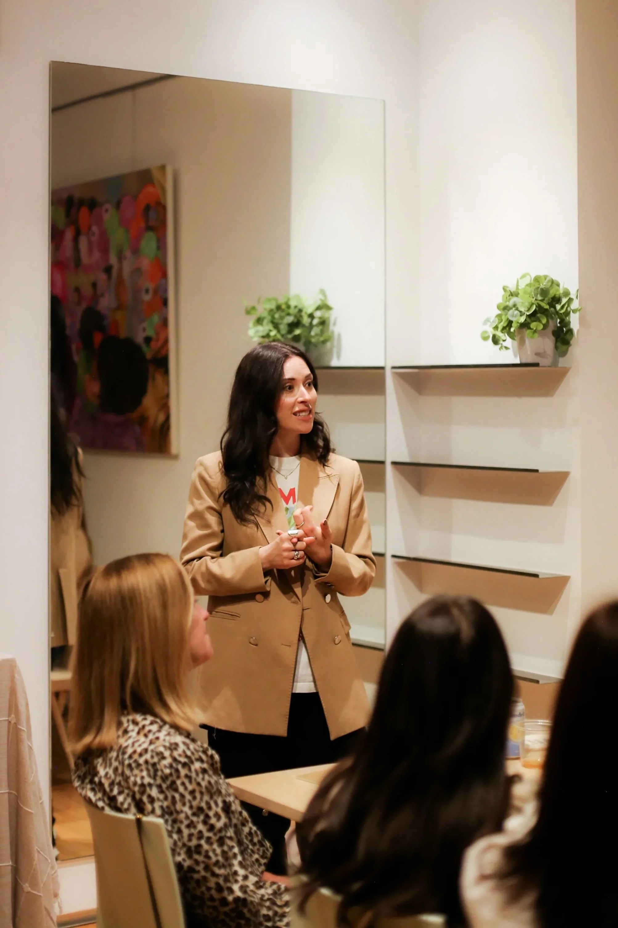 A woman speaking to a group of women seated indoors, with a large mirror, a colorful artwork on the wall, and shelves with plants in the background.