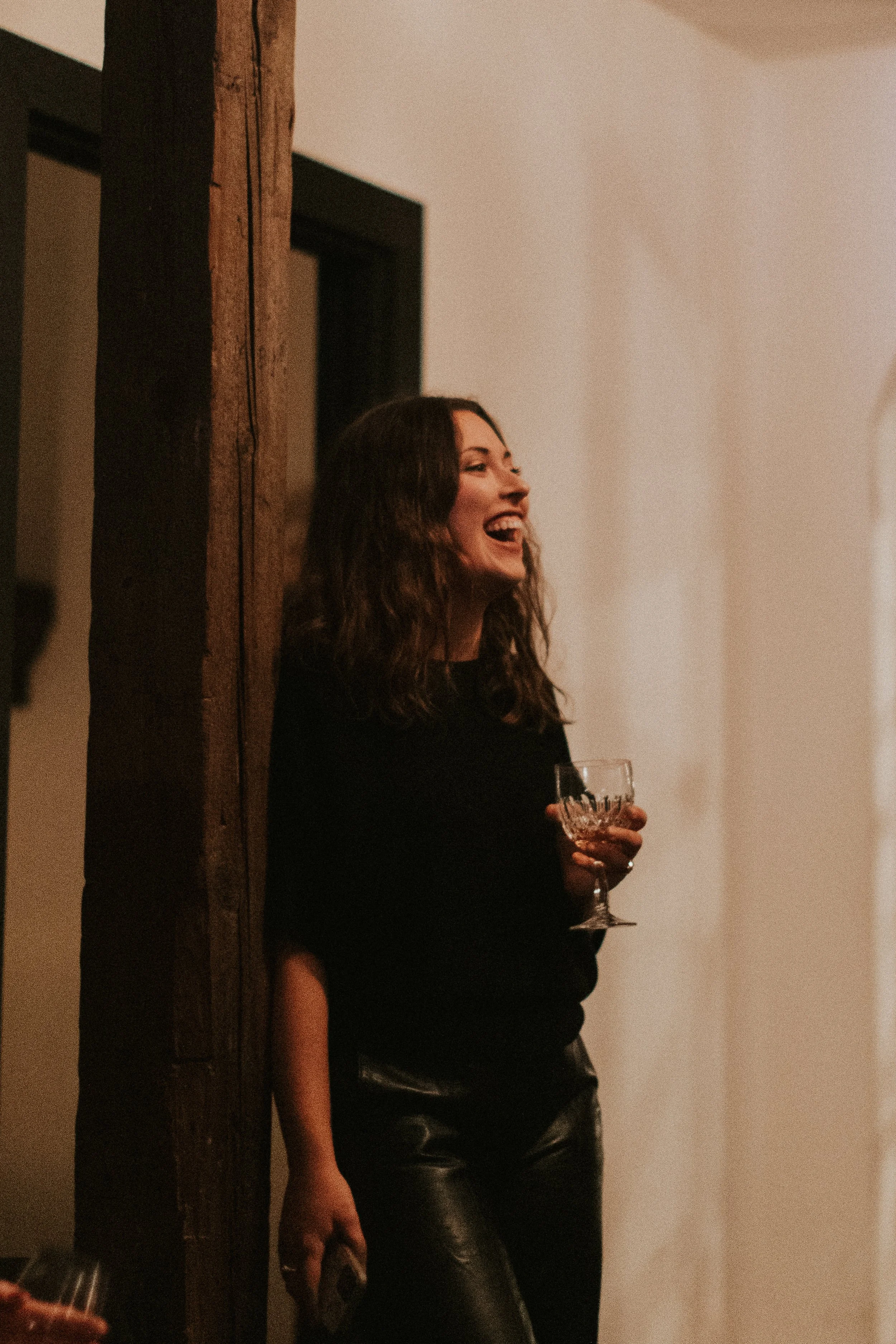 A woman with dark brown curly hair, wearing a black long-sleeve top and black leather pants, is holding a glass with a drink in her right hand, smiling and looking to her right. She is standing against a light-colored wall, partially obscured by a wooden beam.