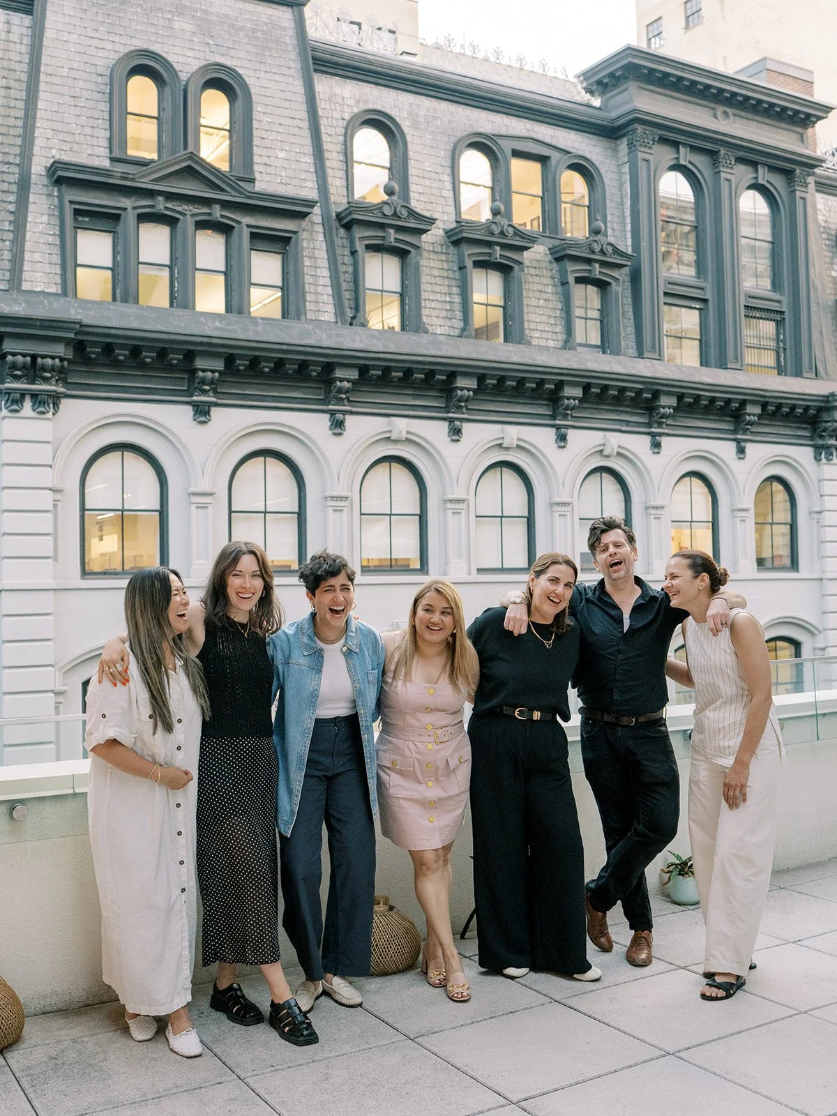 Group of seven diverse women and one man standing together on a rooftop terrace, smiling and laughing, with a historic building in the background.