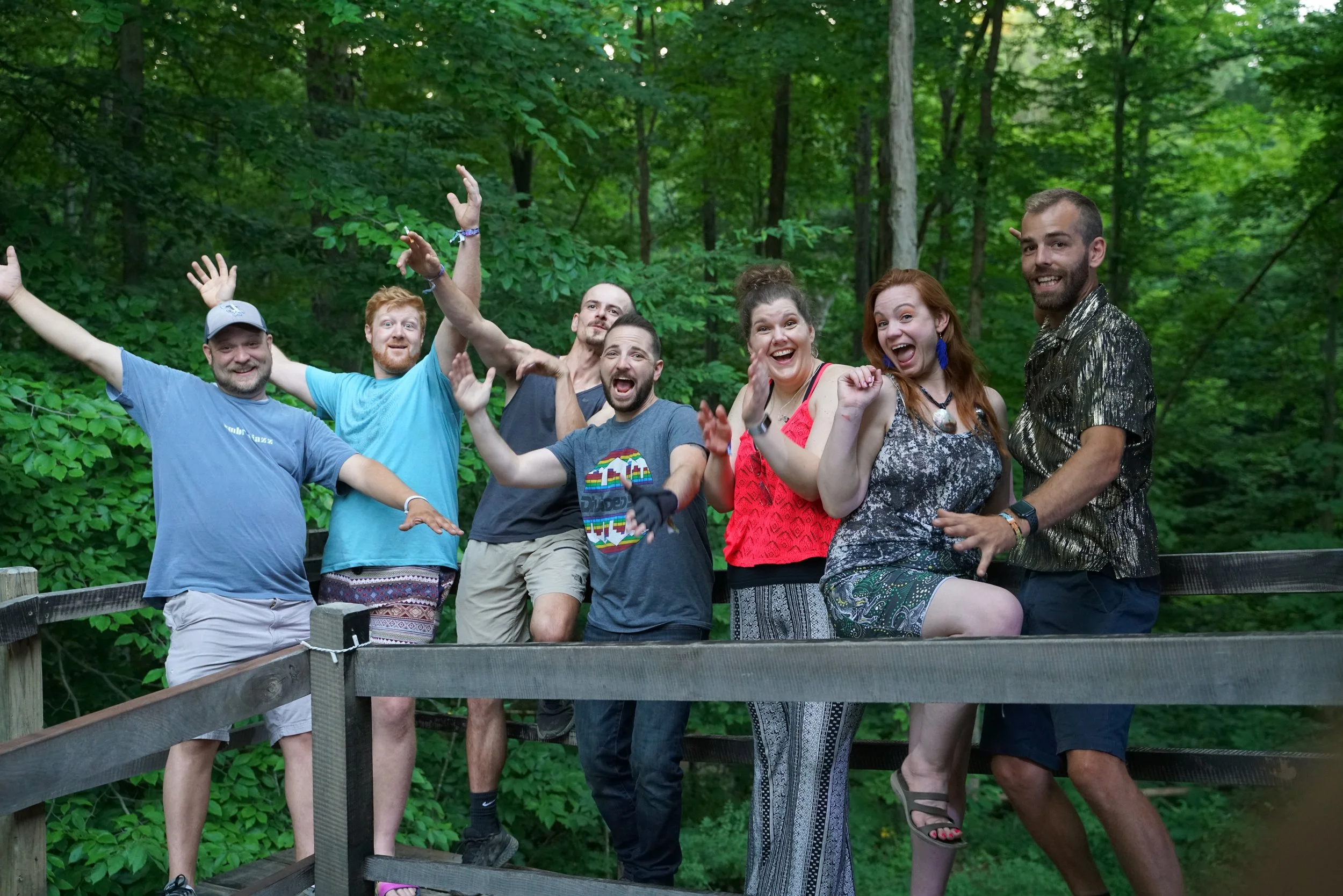 Group of seven people posing playfully on a wooden deck with a forest background.