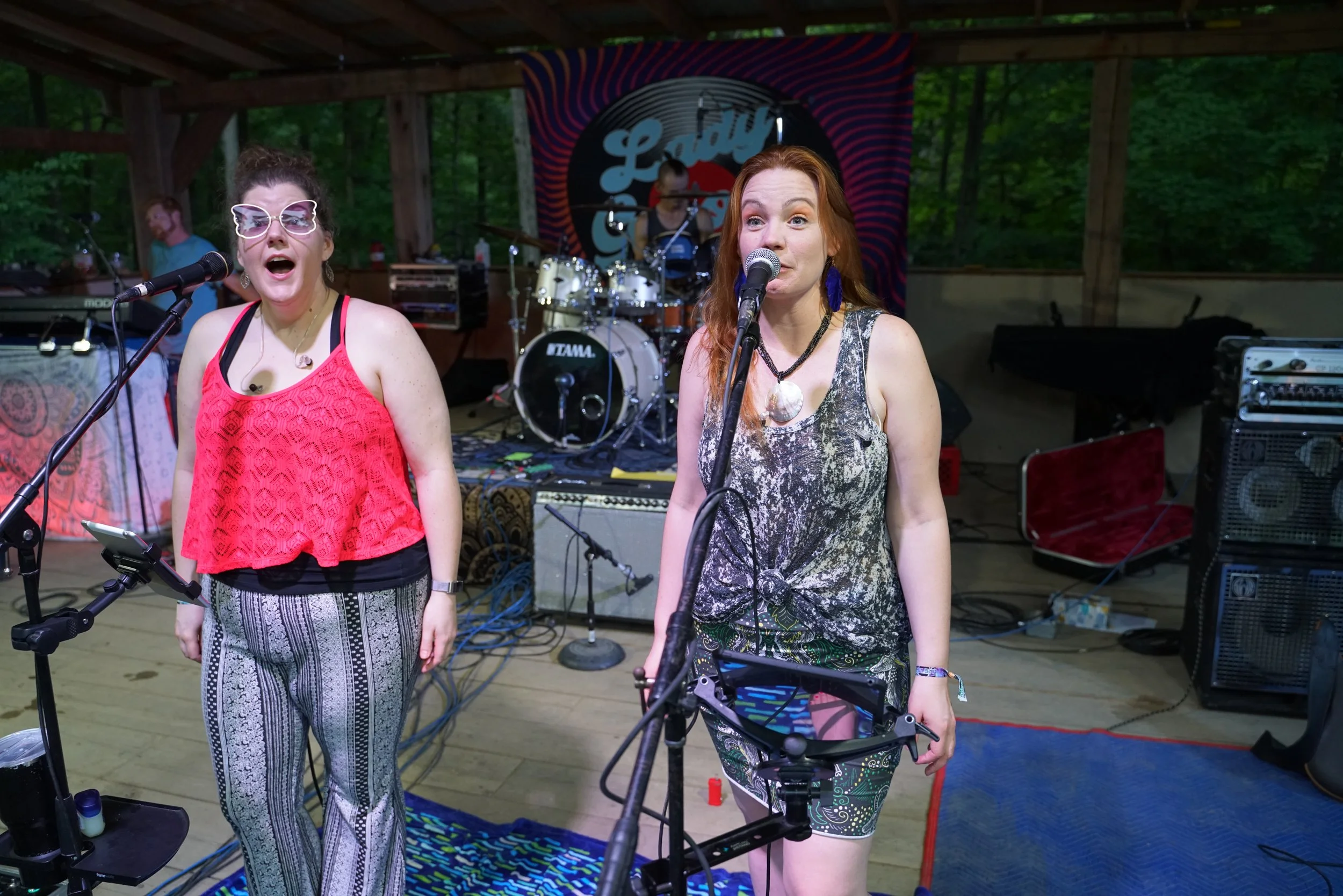 Two women singing on stage at a live outdoor event, surrounded by musical equipment, with a drummer in the background and a "Lady" sign behind them.