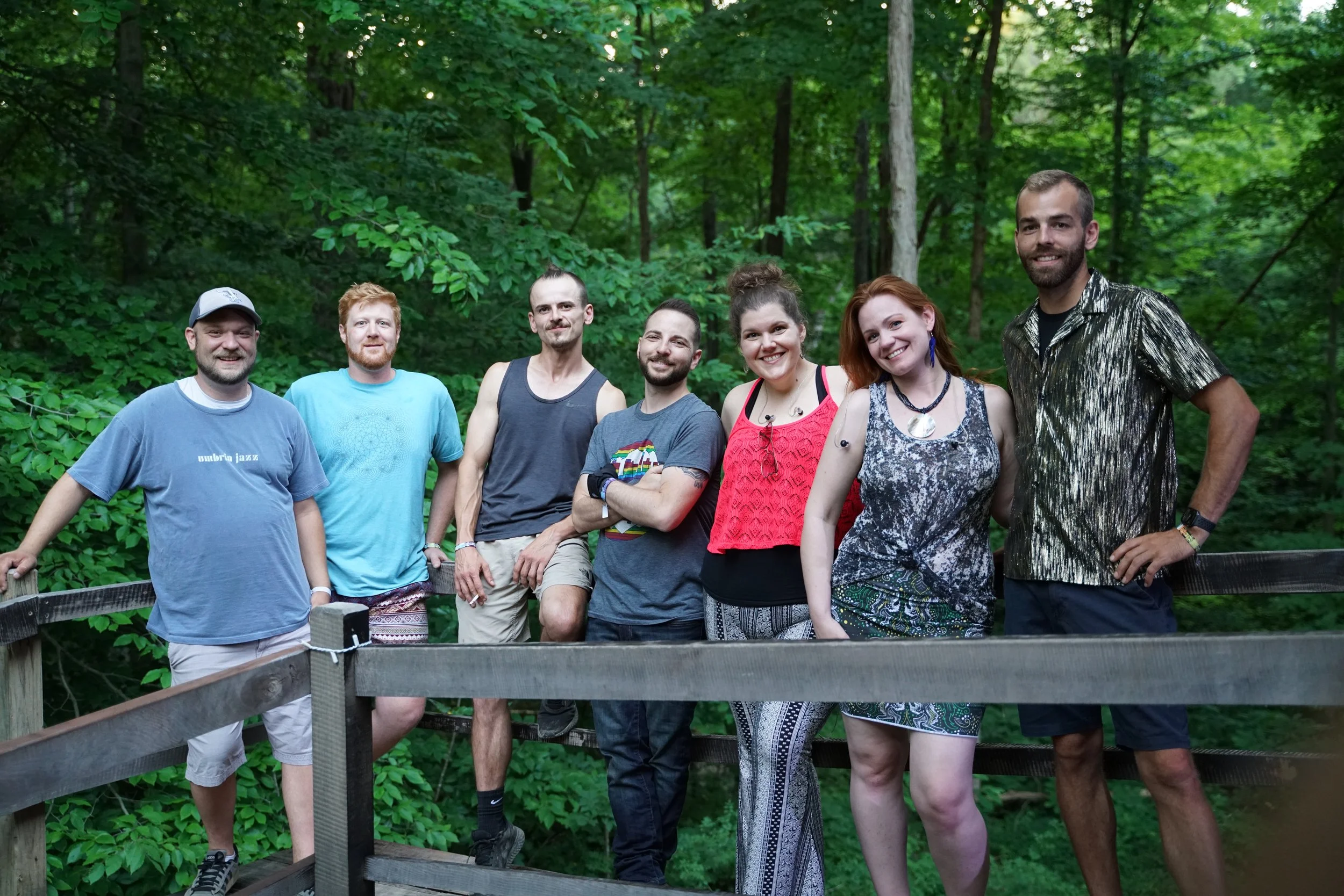 Group of six people posing on a wooden platform surrounded by green forest foliage.
